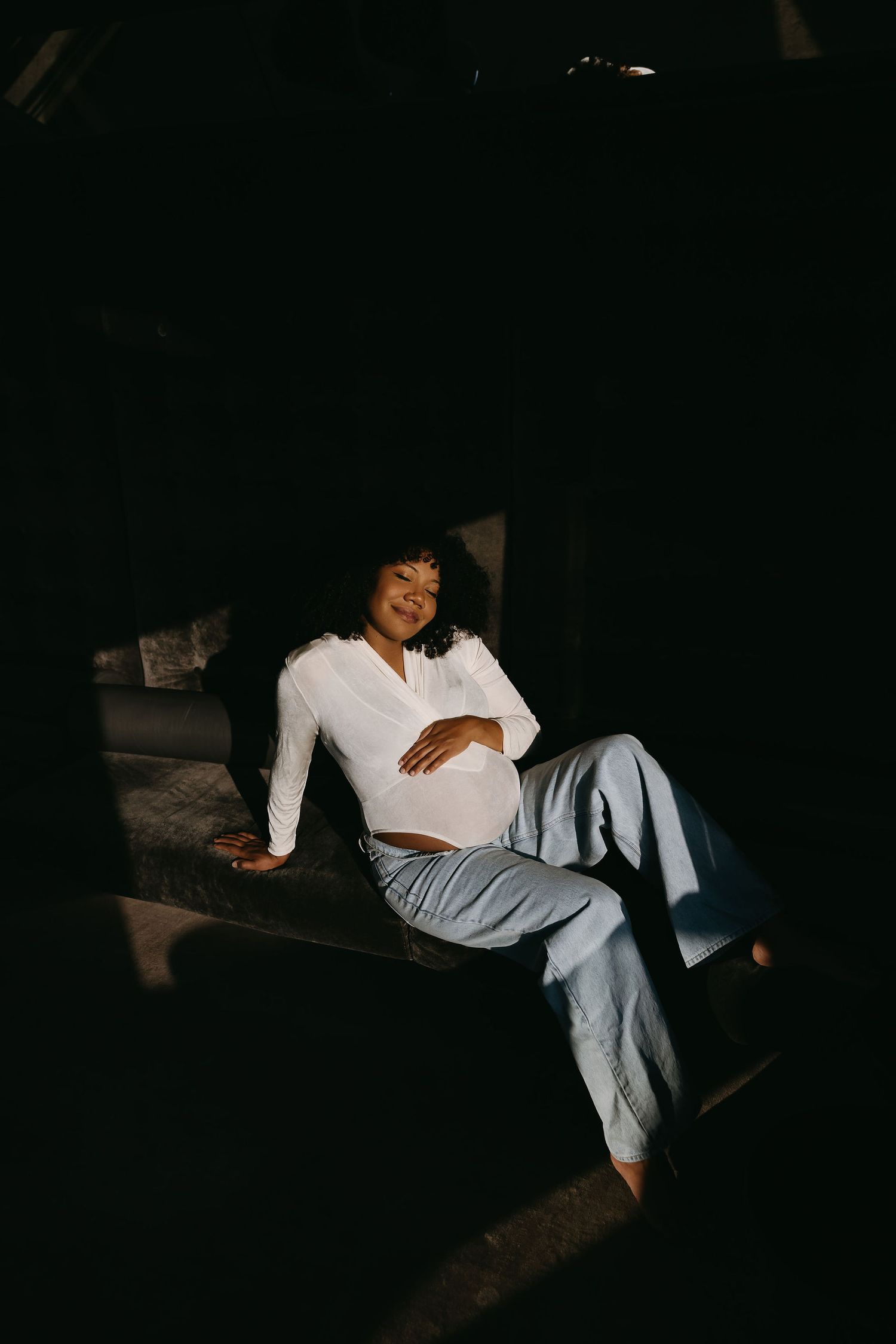 Woman in white loungewear relaxing in shadows on couch during moody lifestyle photoshoot.