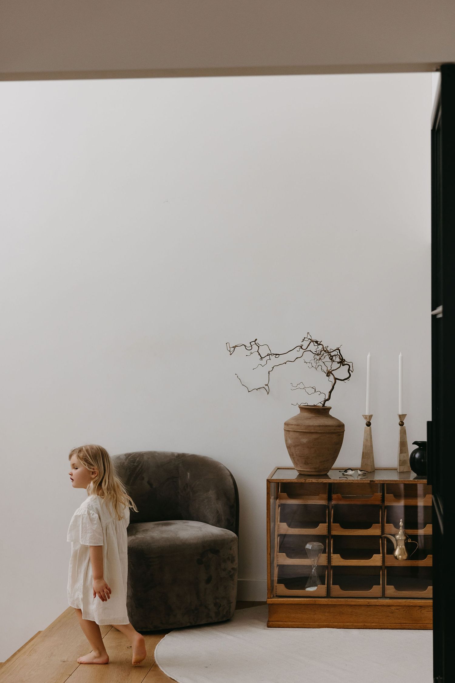 A minimalist interior scene with a child standing near a brown armchair and wooden cabinet.