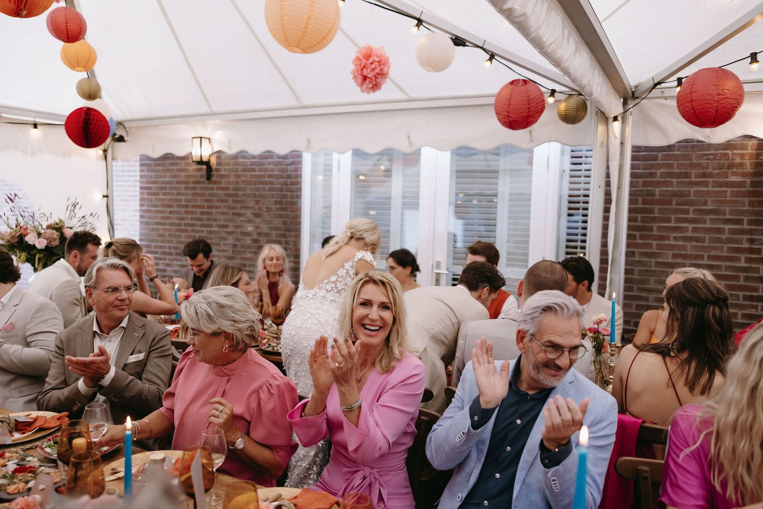 Reception celebration under a tent with pink lanterns and joyful guests in light-colored attire.