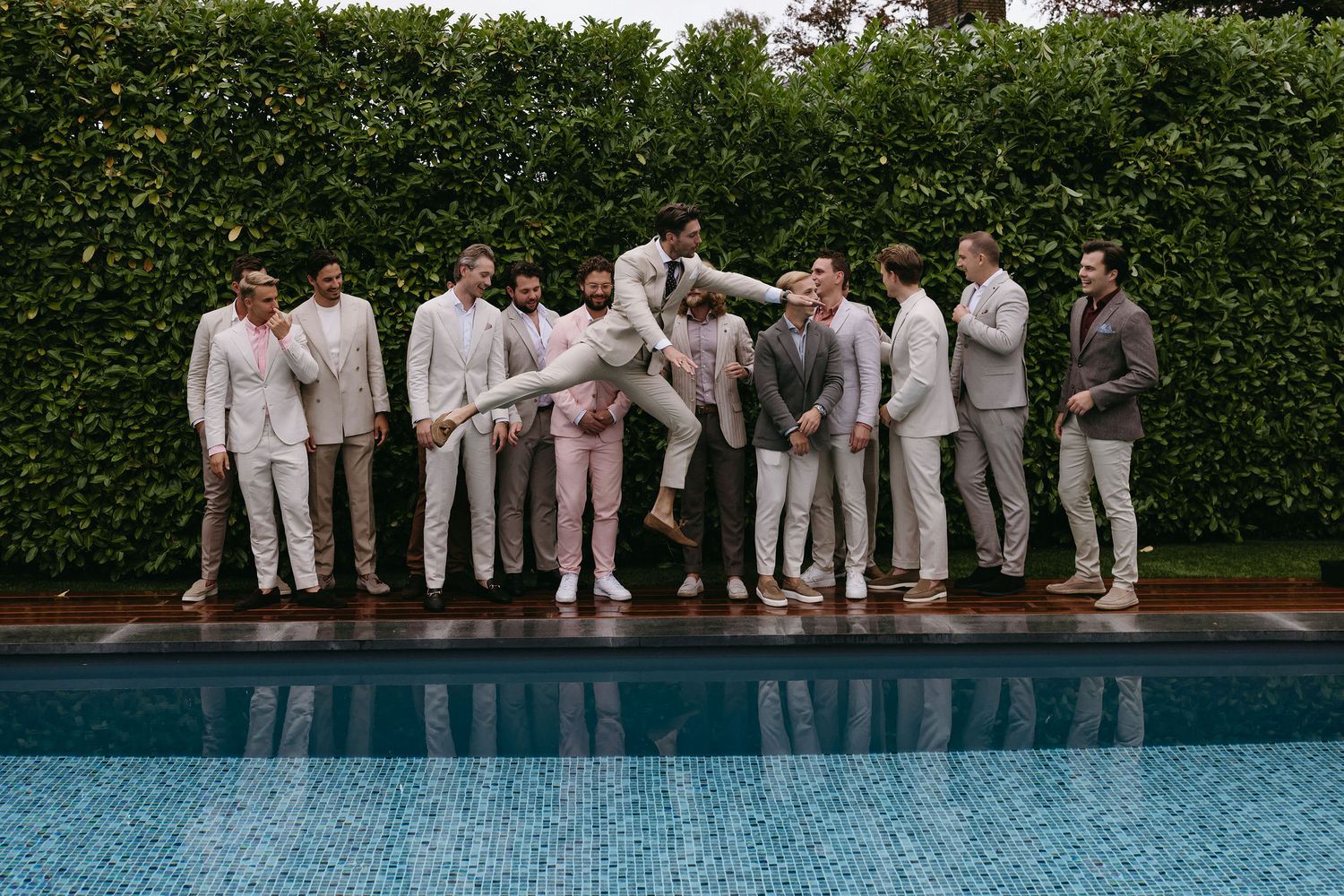 Wedding party in white attire standing along a pool edge with green hedges in background.