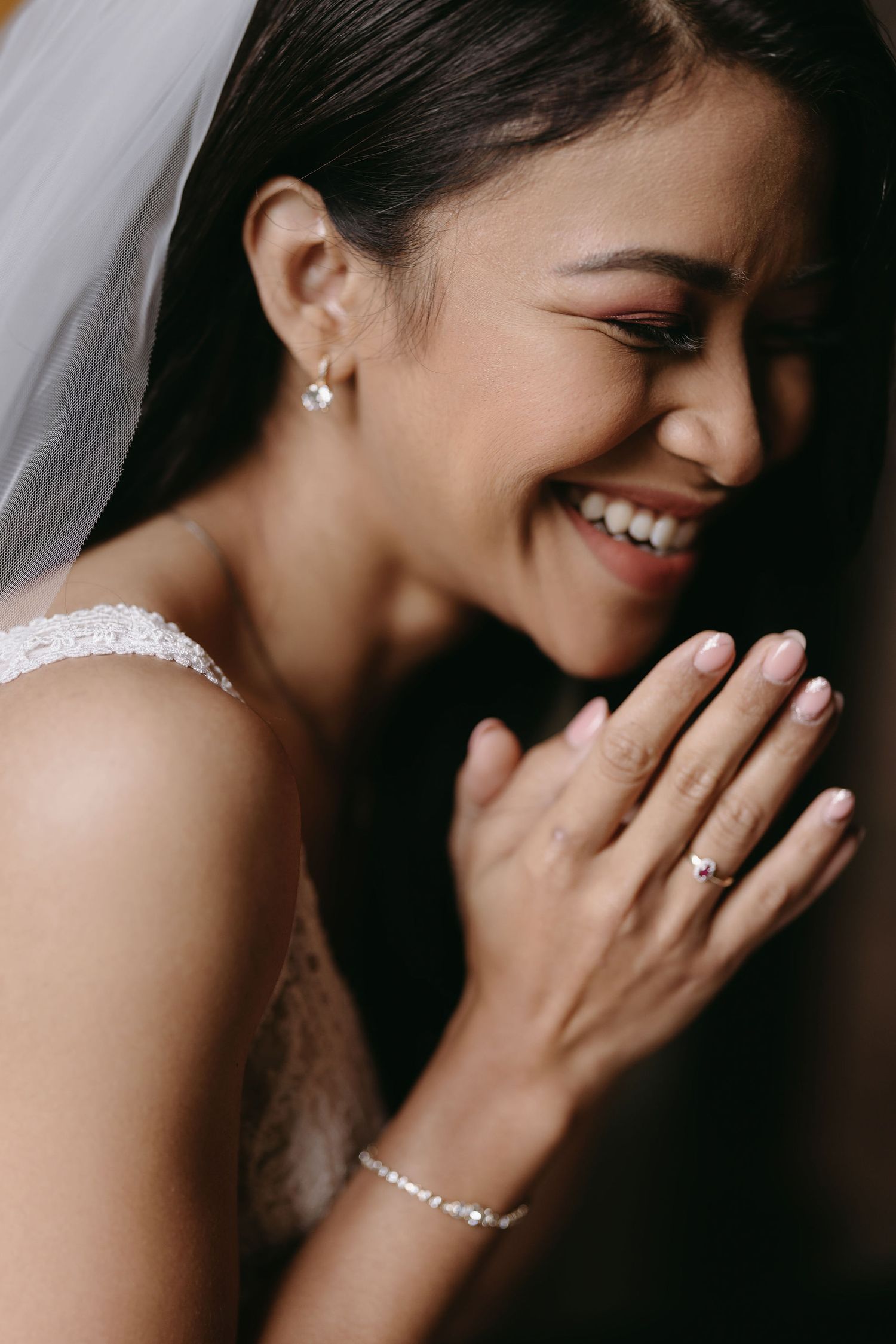 A joyful bride wearing a white lace dress, veil and delicate jewelry smiles with clasped hands in an emotional moment.