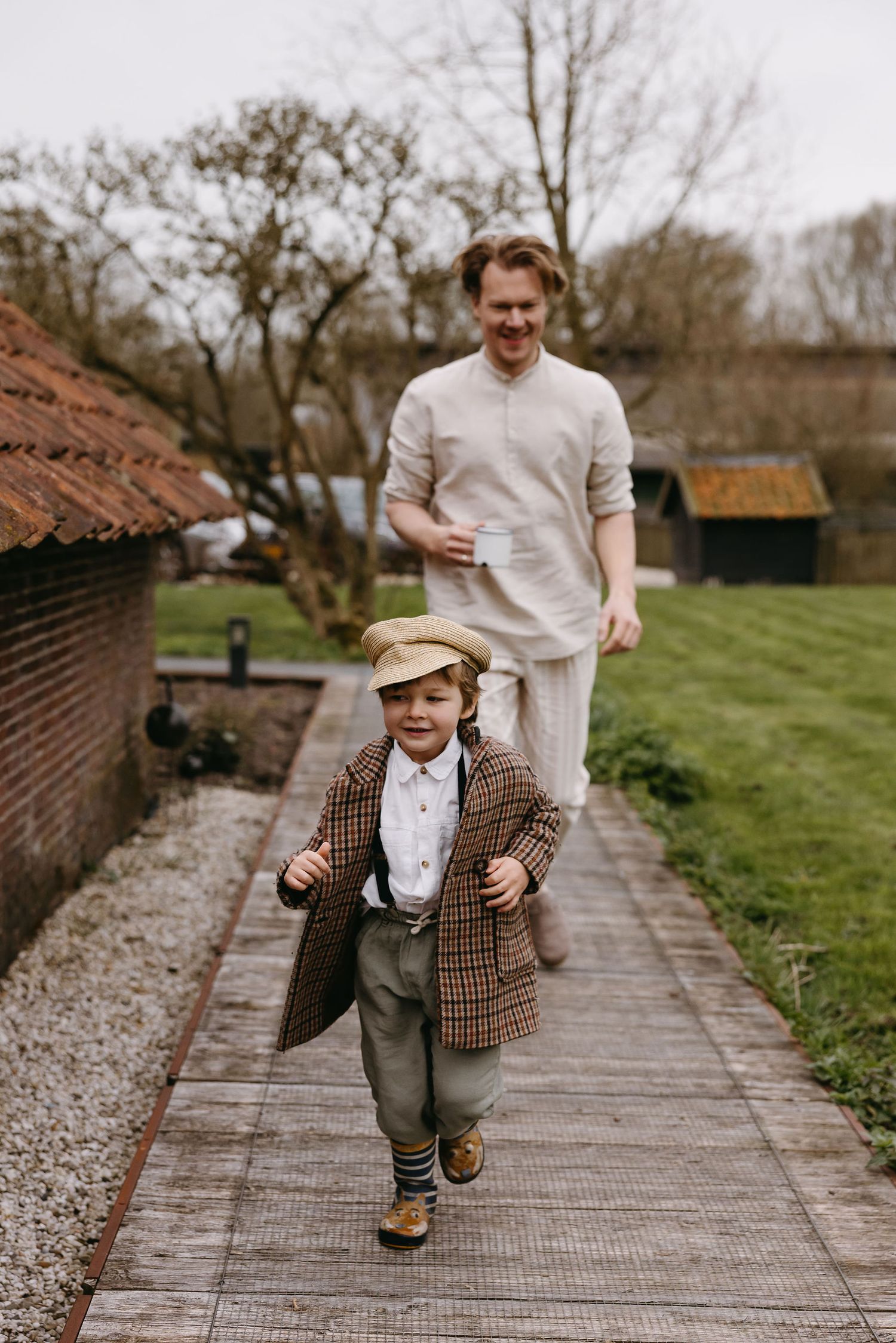 A toddler in vintage-style clothing and cap runs along a garden path while someone follows behind.