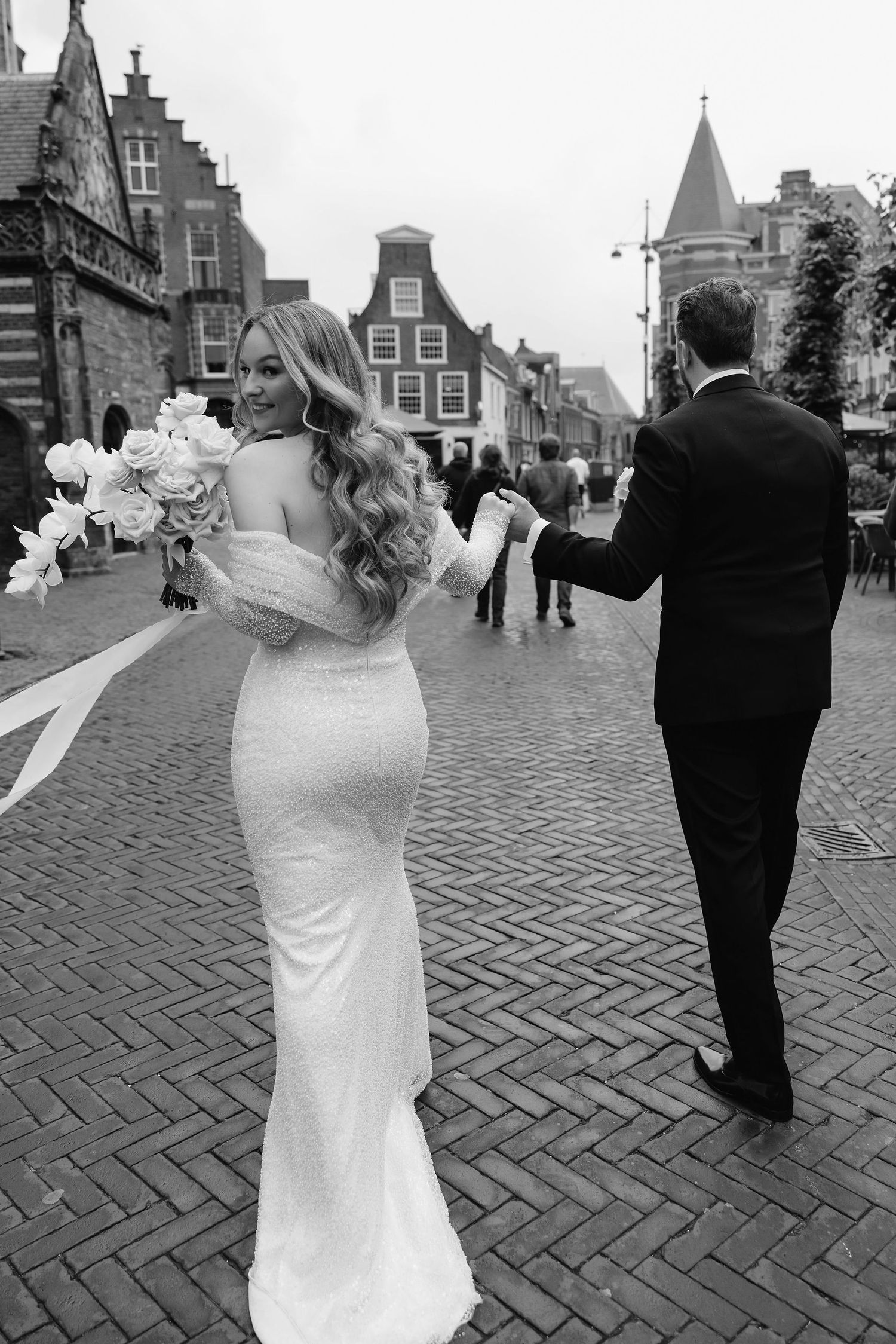 A wedding couple walking on brick streets of a historic European town with traditional Dutch-style buildings in the background.
