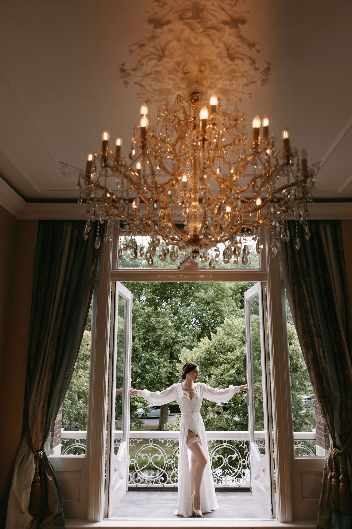 An elegant white gown silhouetted against french doors beneath an ornate crystal chandelier in a luxurious room.