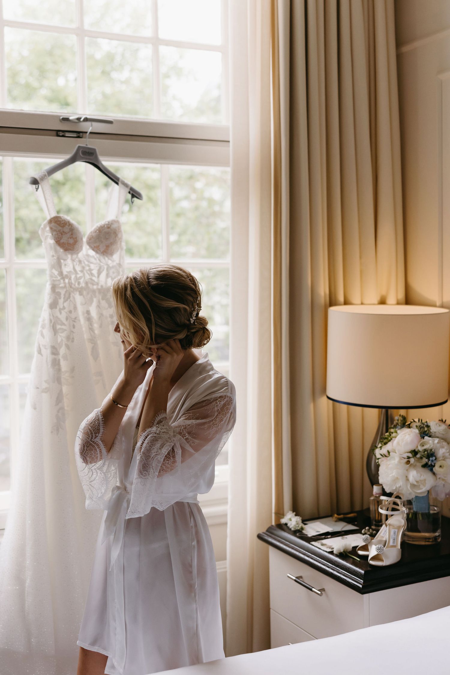 A white wedding dress hangs by a window while someone in a robe looks at it during wedding preparations.