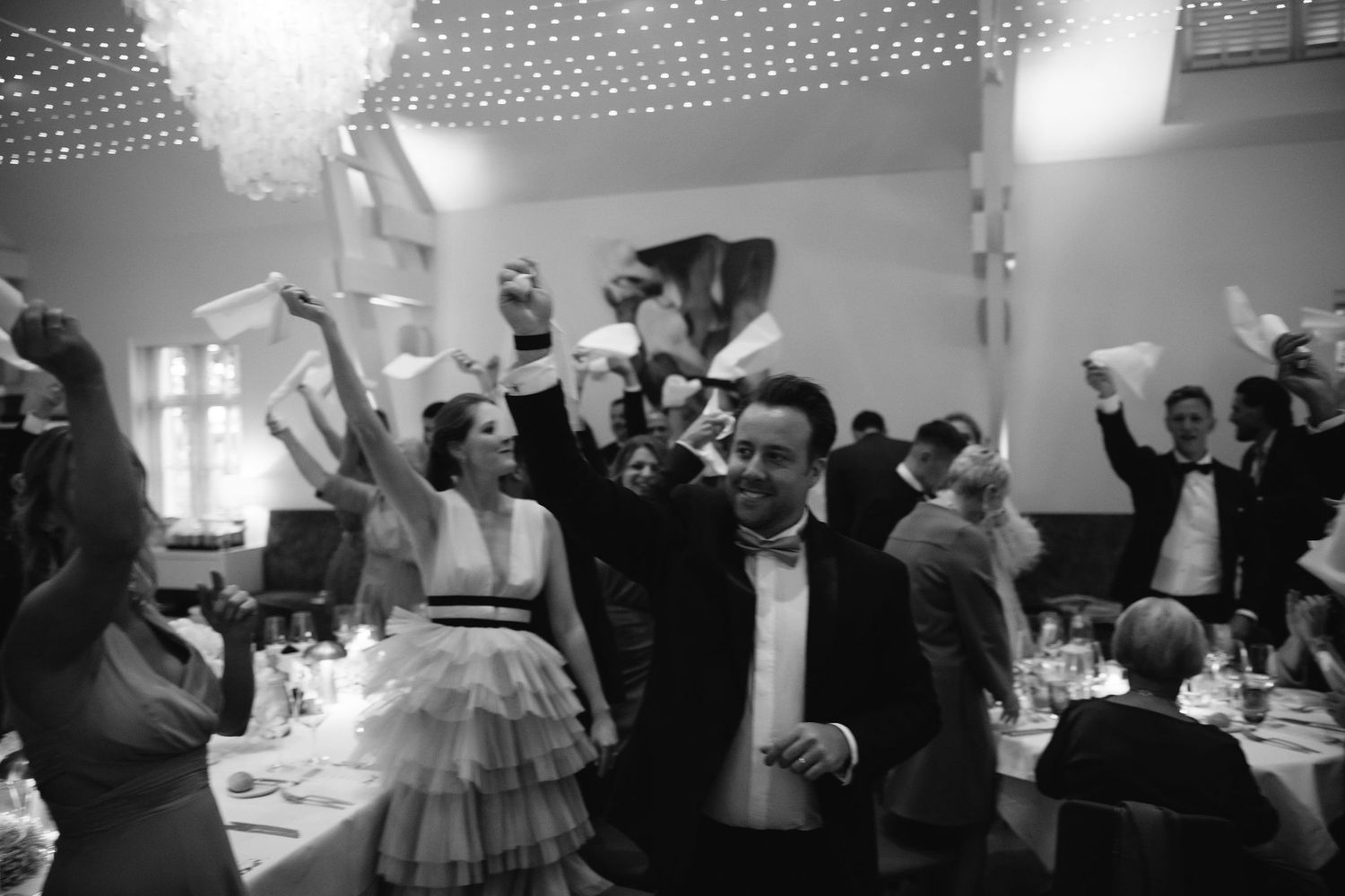 Wedding reception guests dancing and celebrating with raised arms in a festively lit venue.