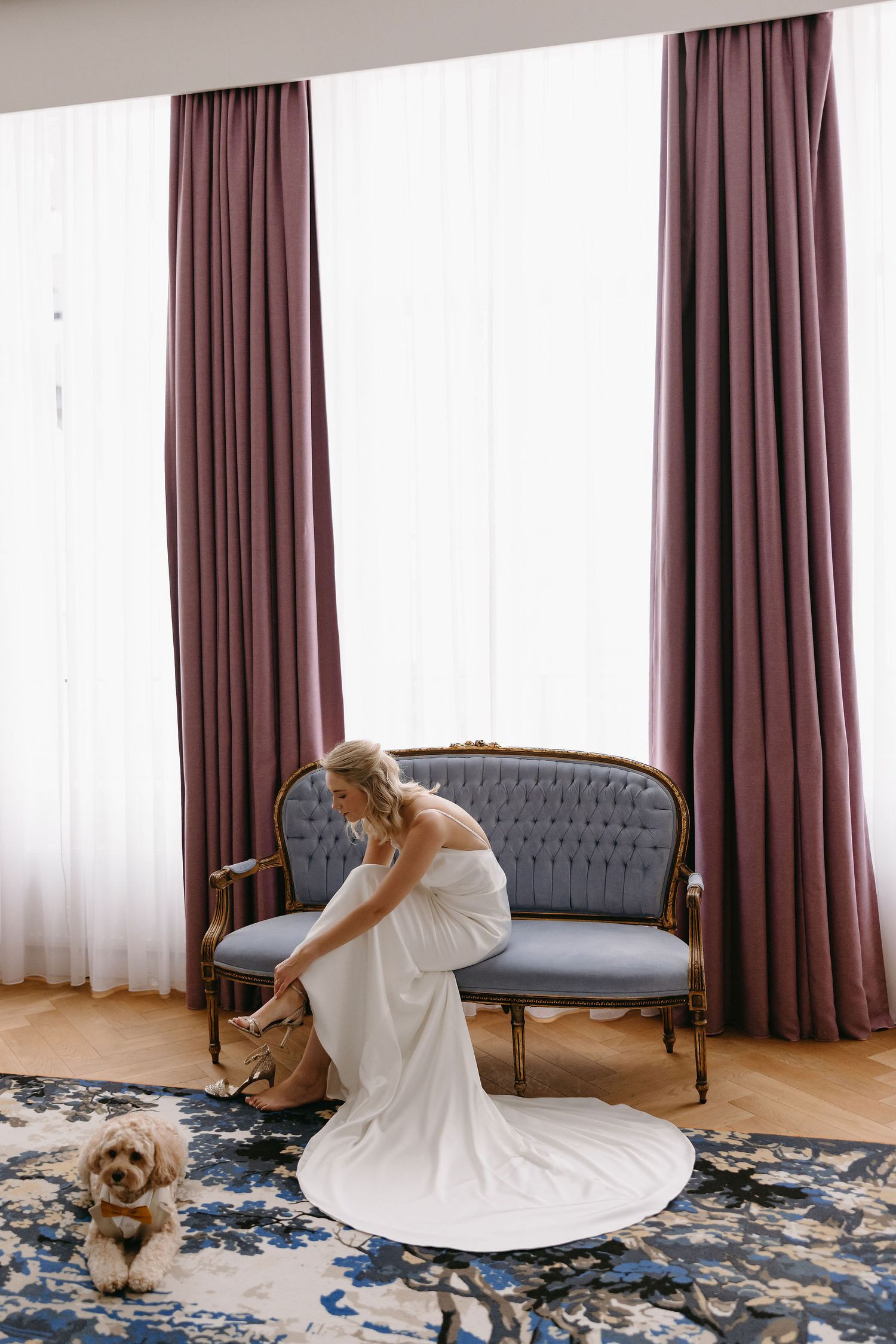 Bride in flowing white dress adjusting shoe on vintage sofa with small dog nearby.