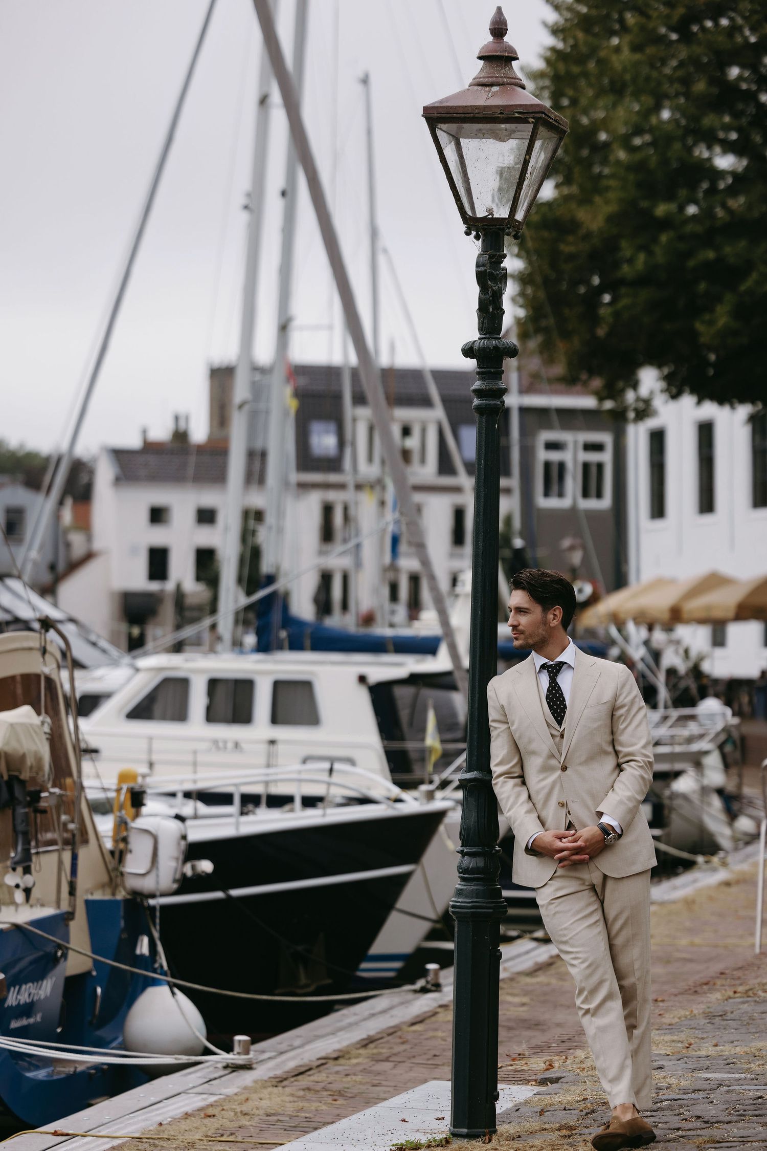 Person in light colored suit stands by vintage lamppost at marina with boats in background.