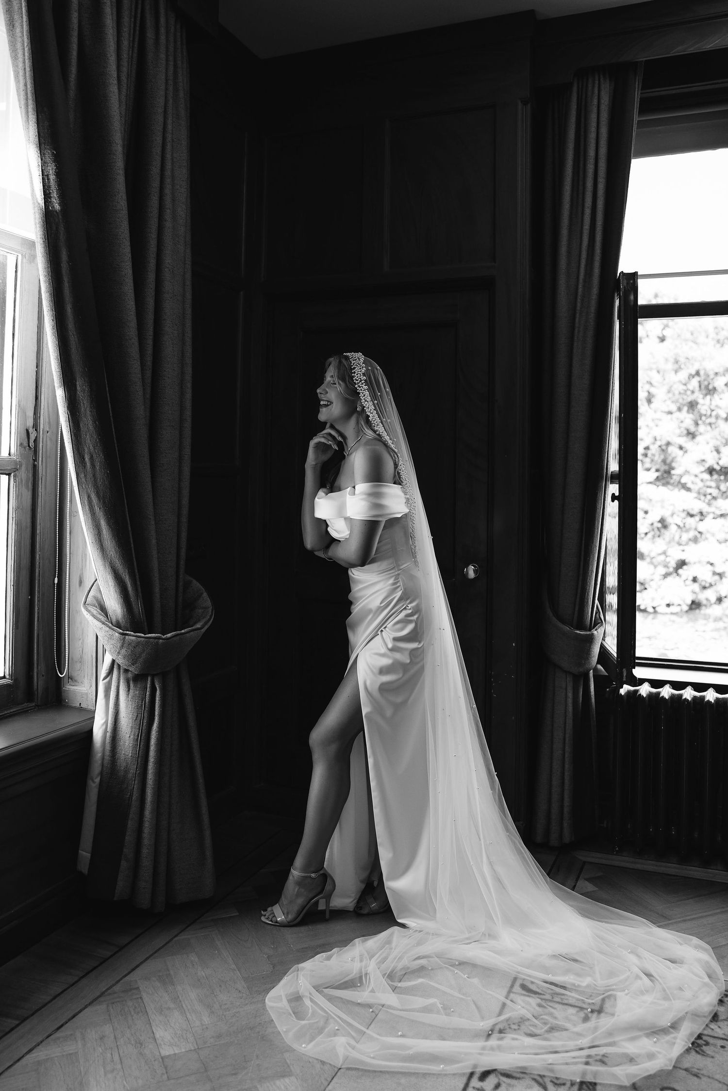 A flowing white wedding dress with a long train is arranged by a window in dramatic black and white lighting.