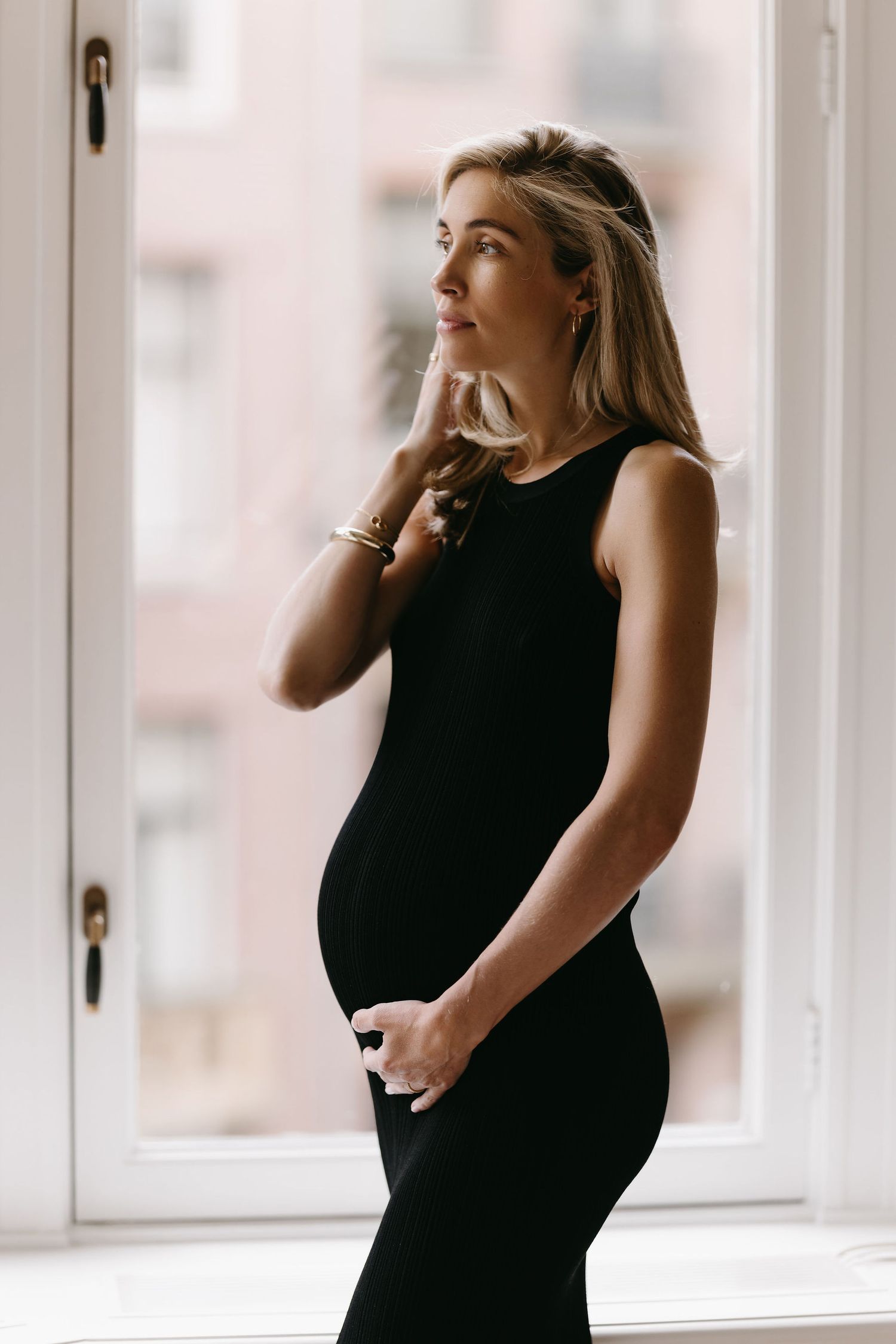 Maternity photo of woman in black dress standing thoughtfully by window.