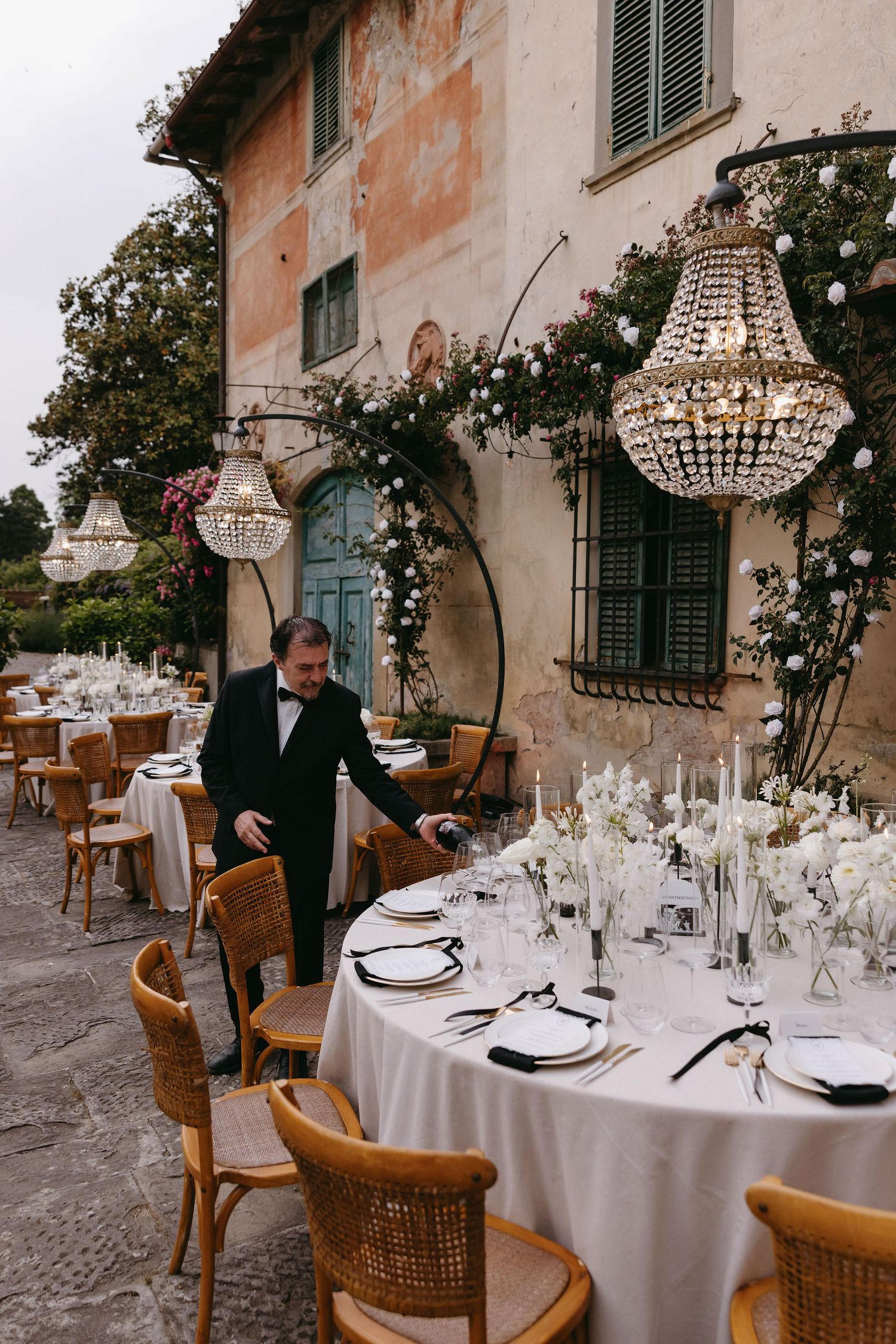 Outdoor wedding reception setup with crystal chandeliers and white tablecloths against a terracotta villa.