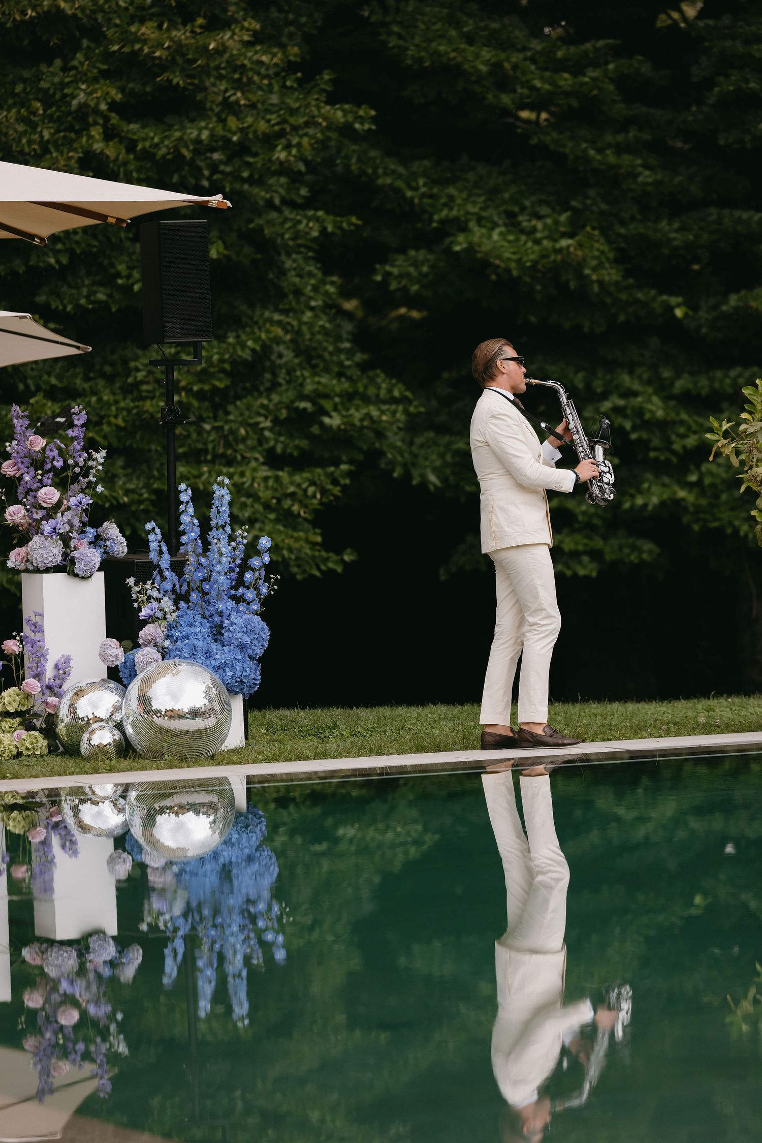 Person in white suit giving speech by reflective pool with purple flowers in garden setting.