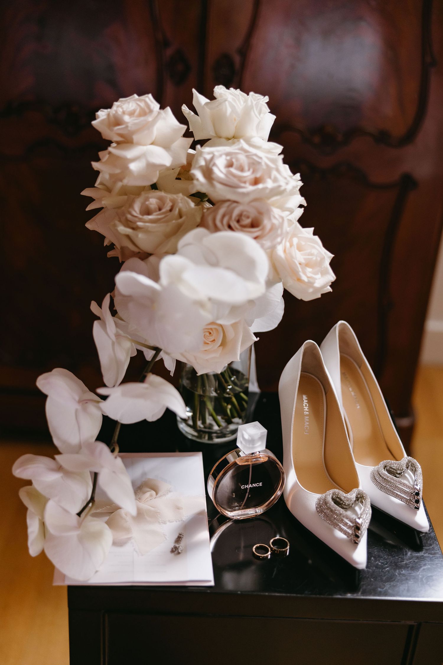 Wedding details including white roses, bridal shoes, perfume bottle and rings on a dark surface.