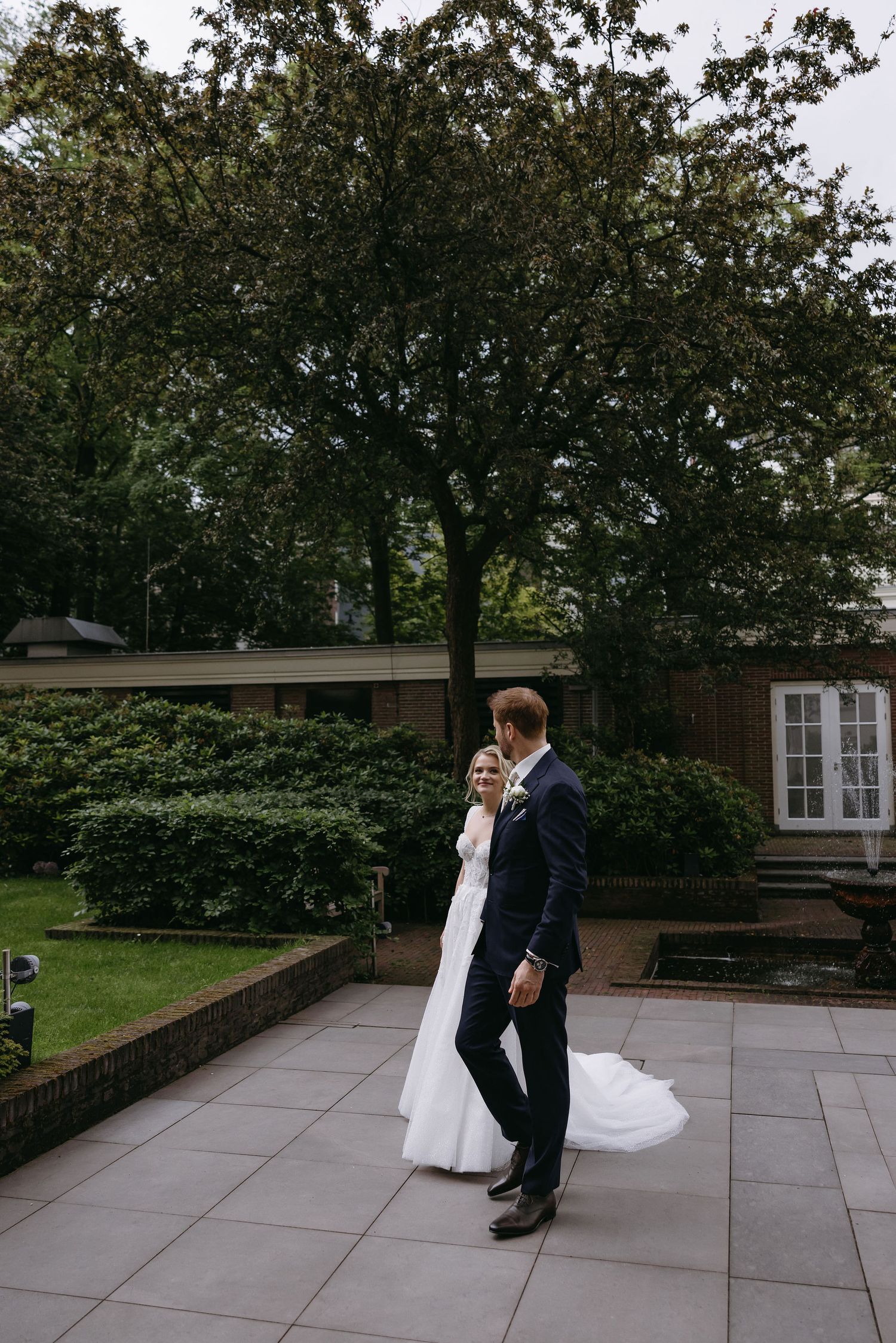 A couple share a romantic moment on a paved garden terrace with lush greenery in the background.