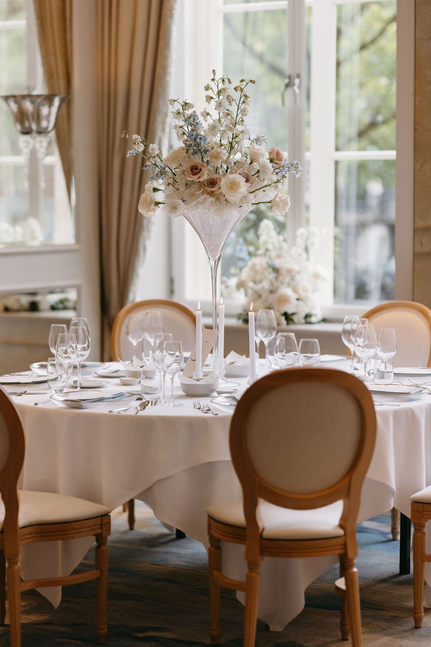 Elegant dining room set with white tablecloths and classic wooden chairs next to bright windows.