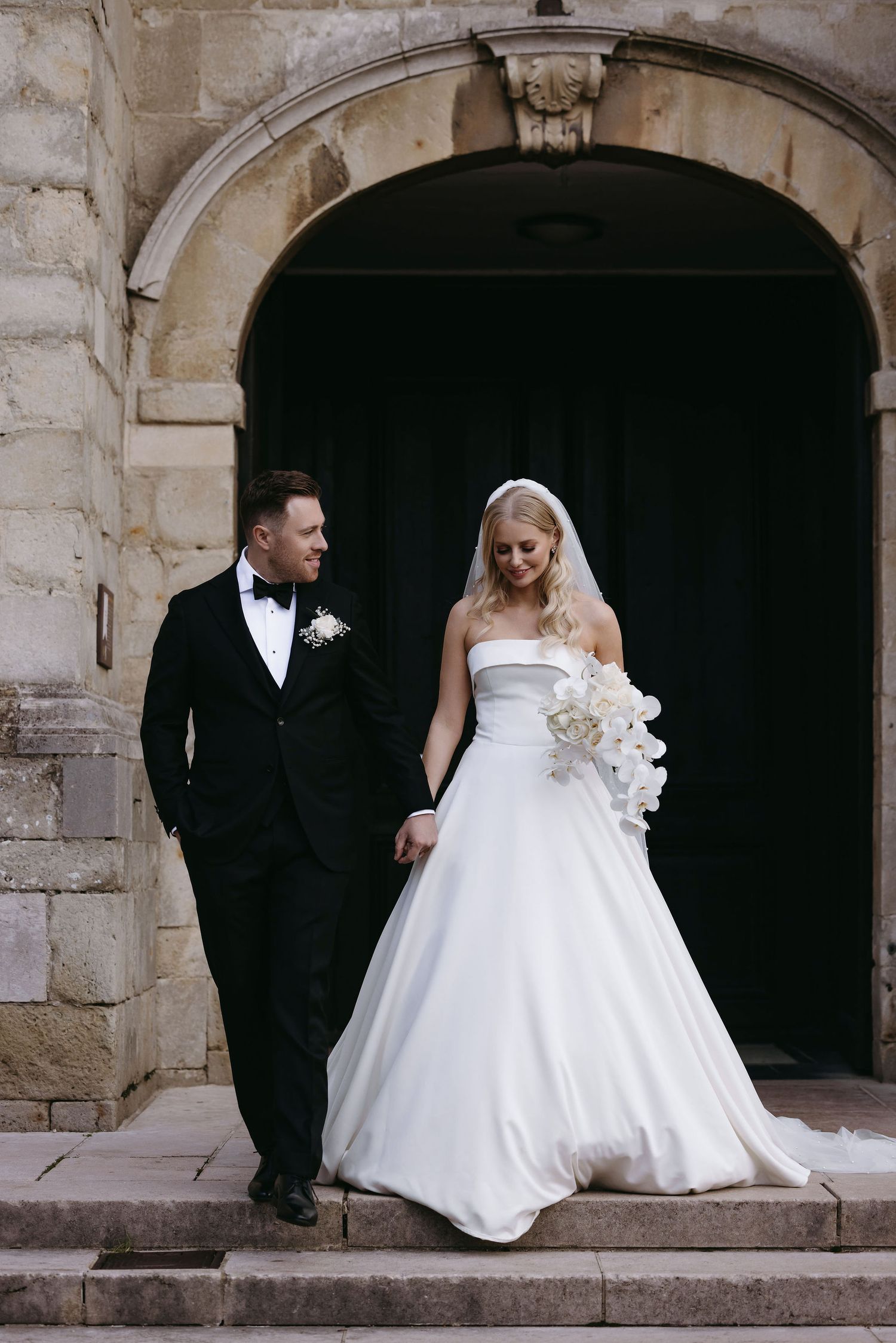 Wedding couple poses under stone archway, bride in classic white gown and groom in black tuxedo.