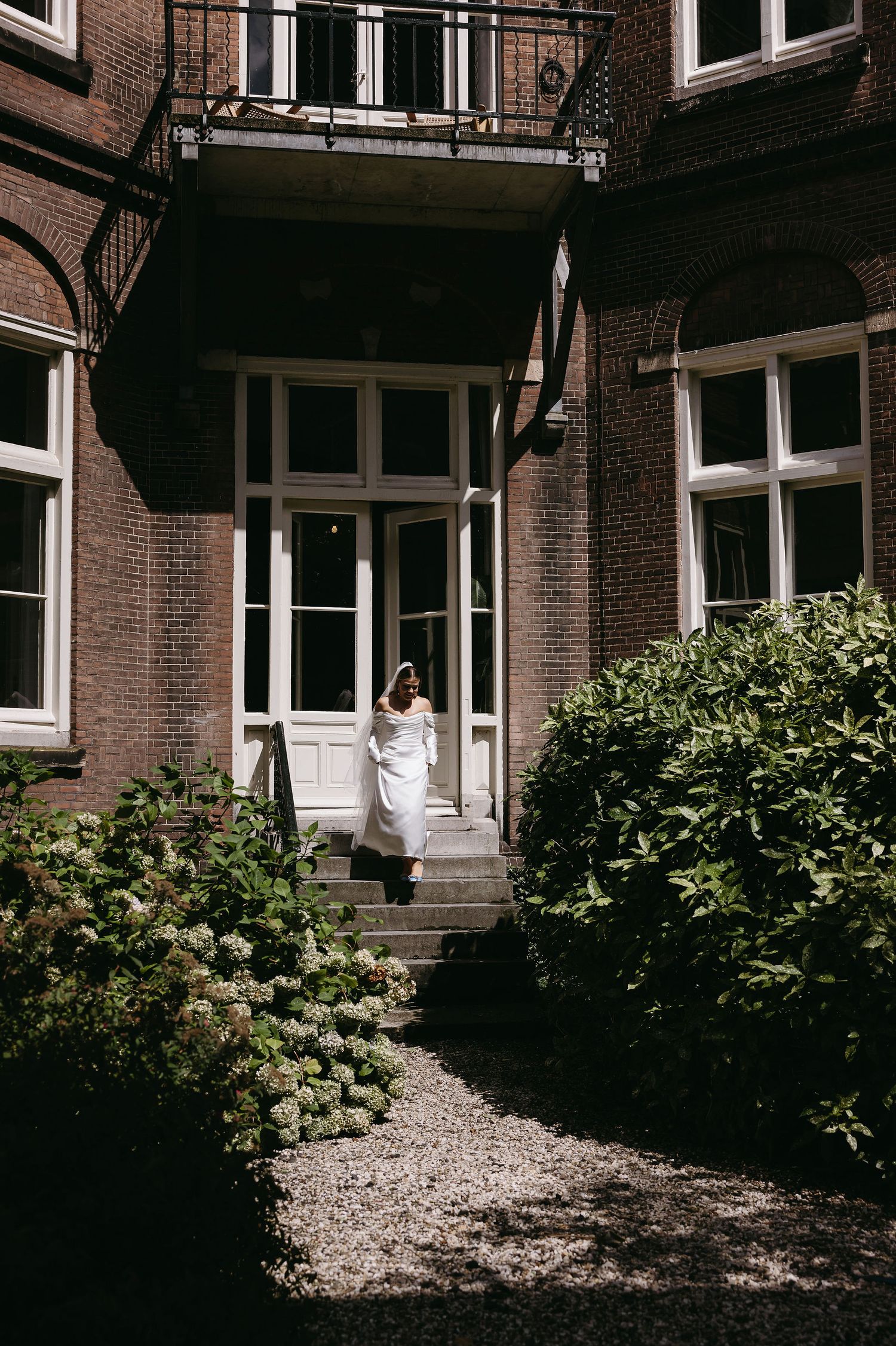 White-clad figure stands on steps of brick building with greenery in foreground.