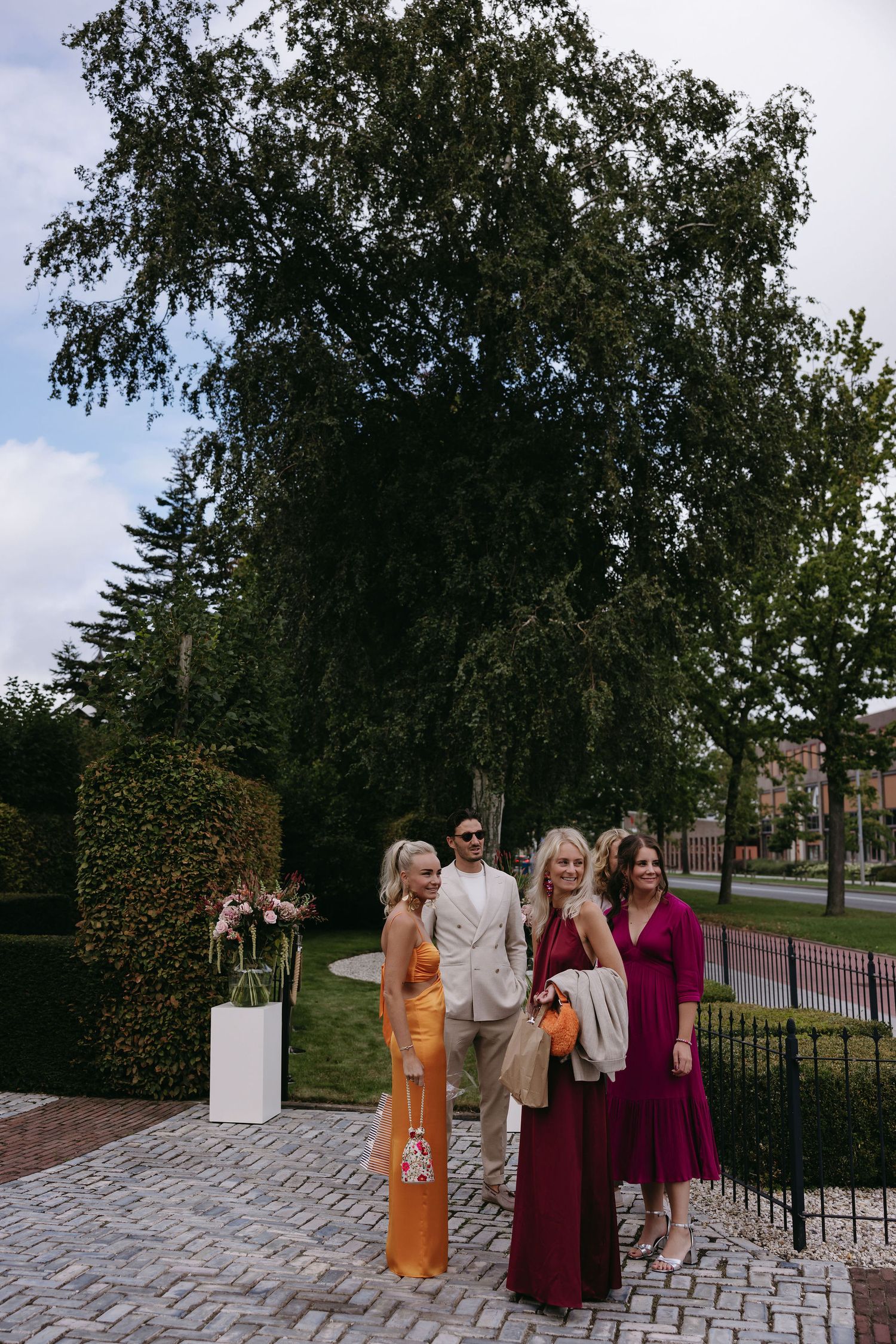 Wedding party poses for photos on a brick pathway surrounded by trees and greenery.