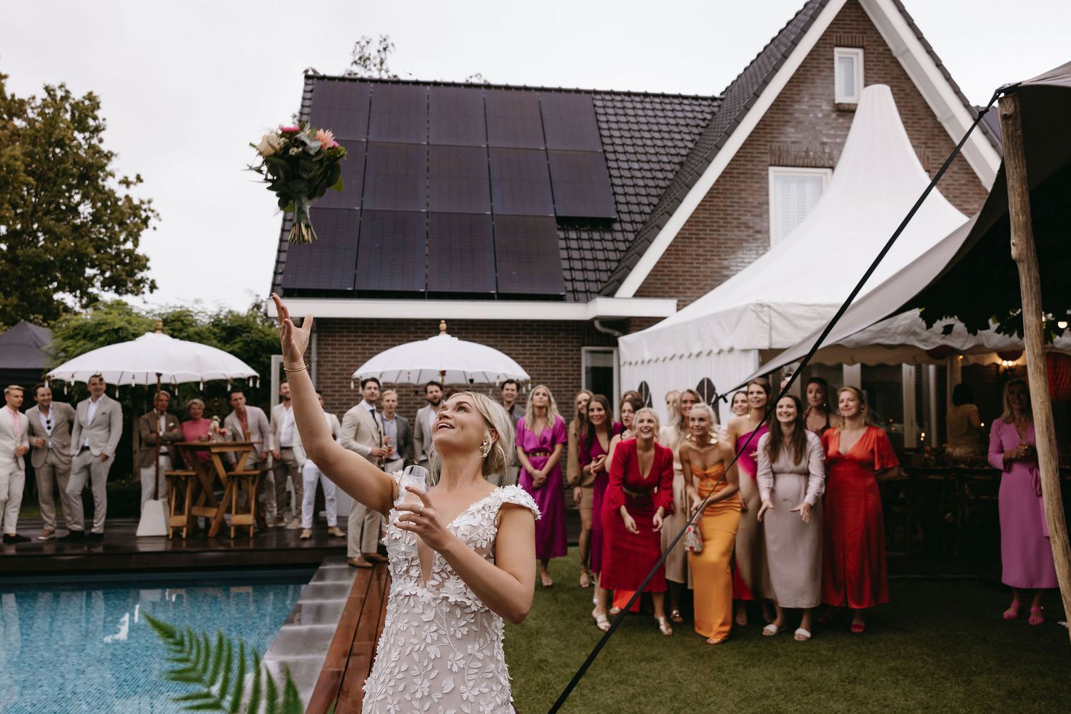 Bride tosses bouquet to waiting guests by poolside at outdoor wedding reception.