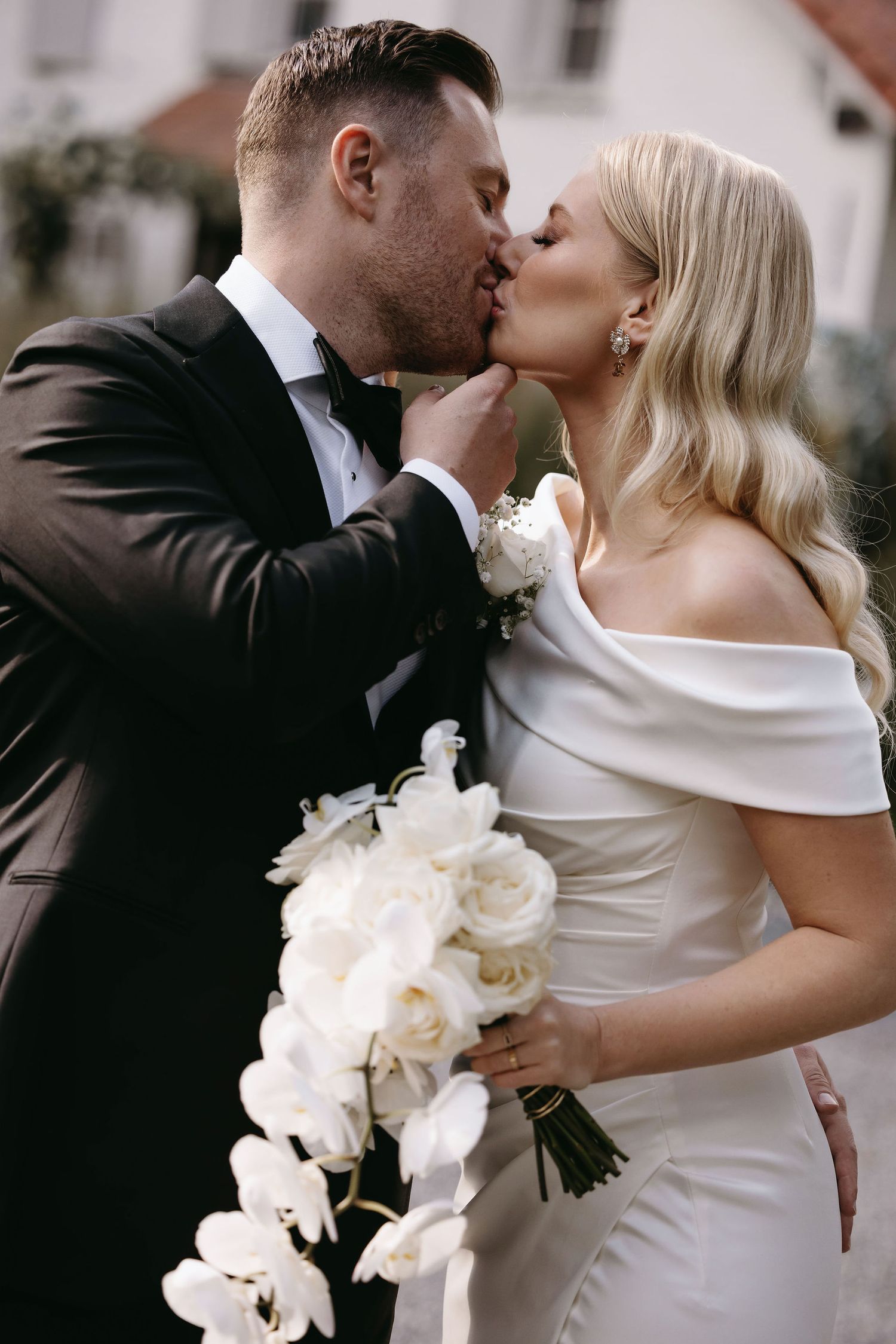 Elegant wedding couple sharing romantic kiss while holding white orchid bouquet.