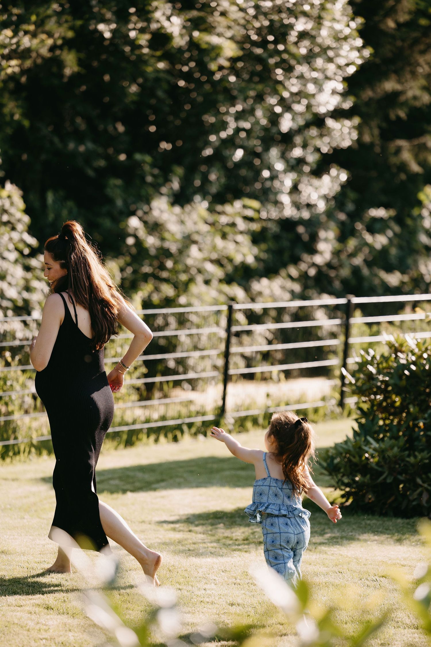 A parent and small child in overalls walking together on a sunny path with nature and railing in the background.
