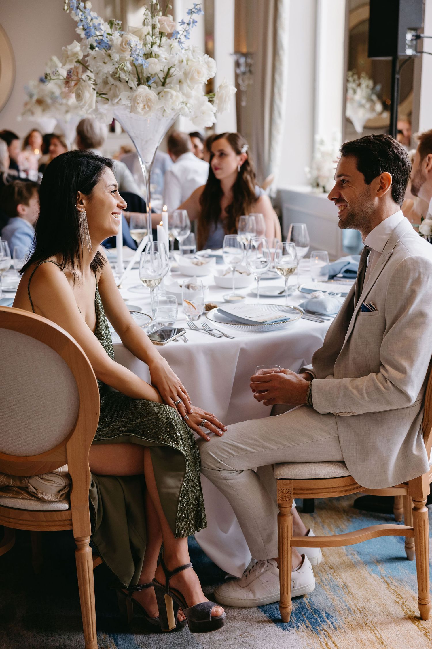 Wedding reception guests sit at elegantly decorated tables with tall floral centerpieces.
