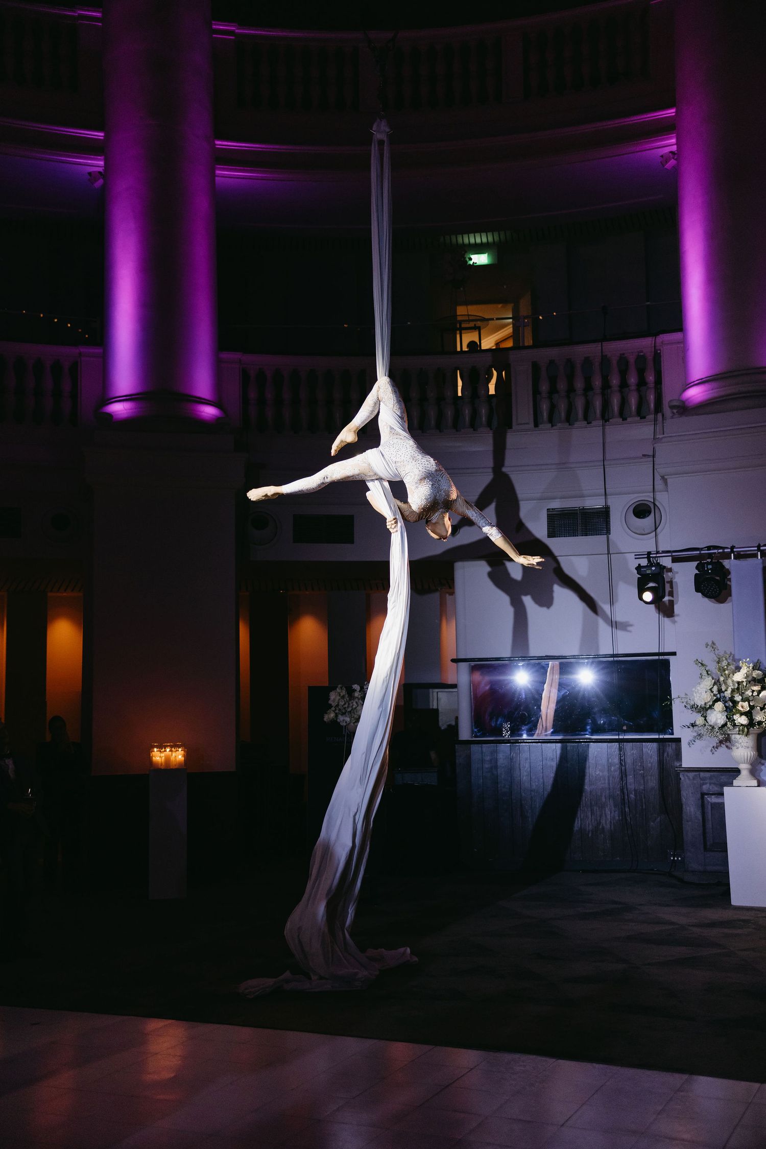 Aerial silk performer executing graceful moves under dramatic purple lighting at an elegant indoor venue.