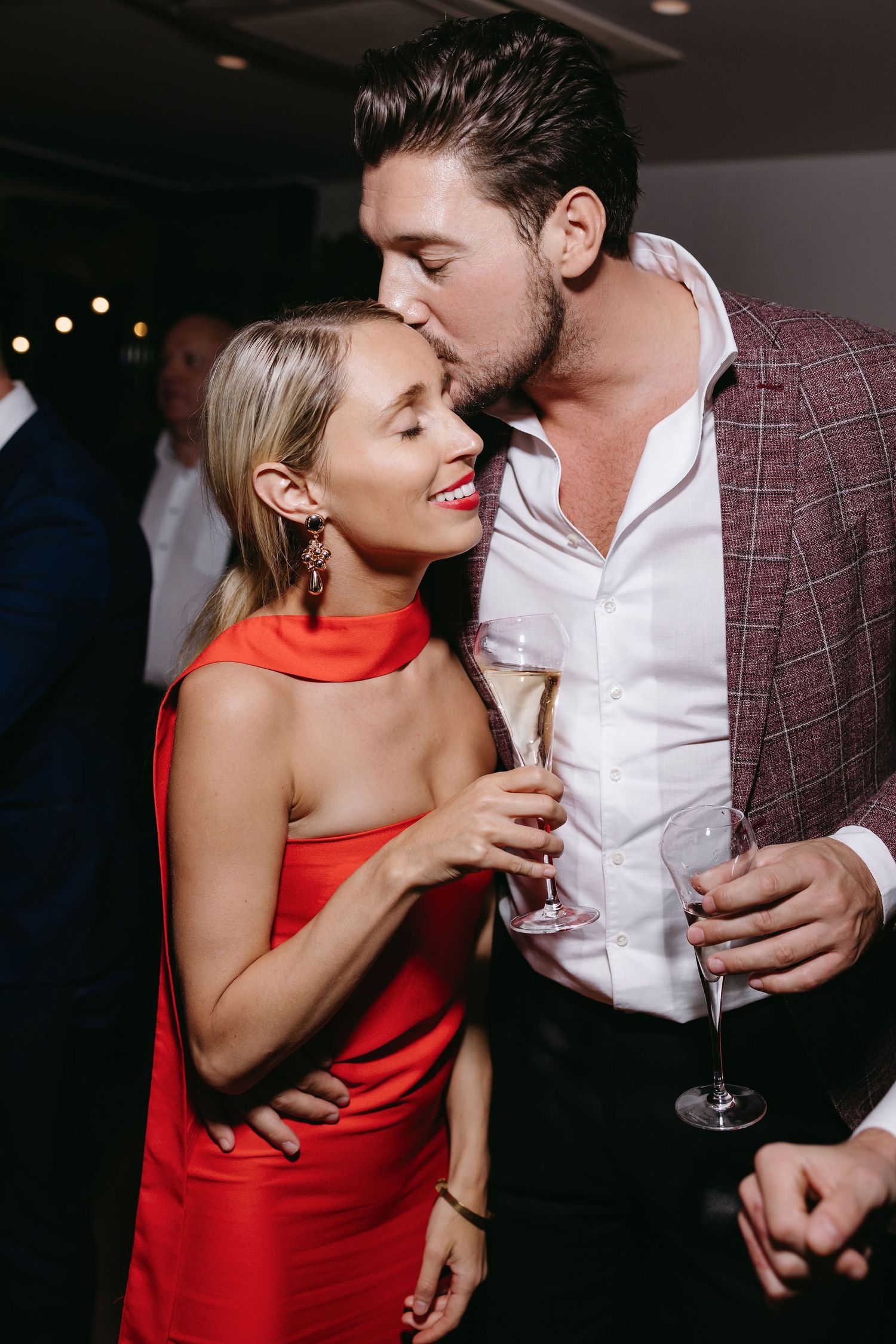 A couple shares an intimate moment at a party, with her in a red dress and him in a plaid shirt holding champagne glasses.