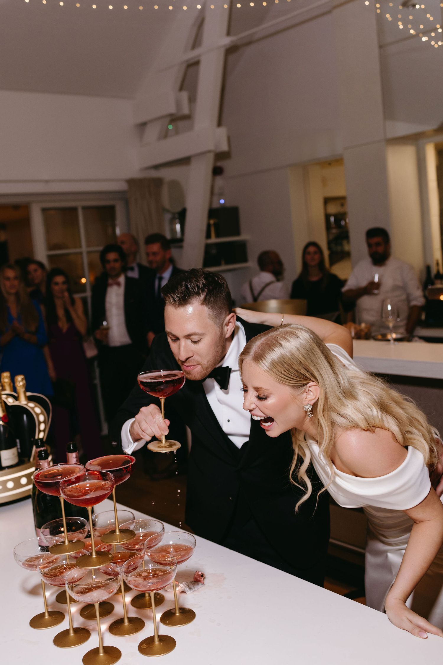 Couple leaning over champagne tower at wedding reception with string lights overhead.
