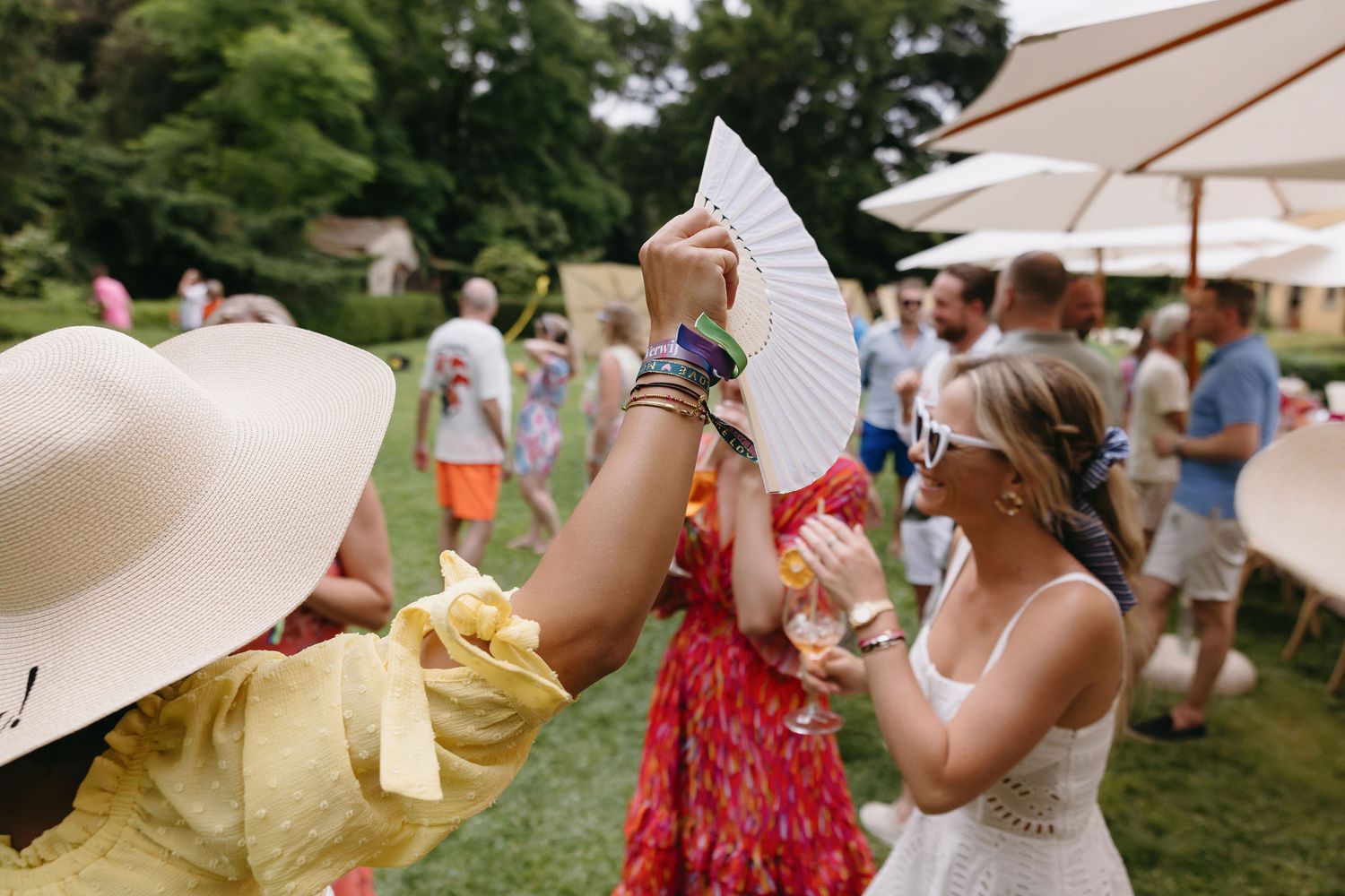 Summer garden party guests fanning themselves with paper parasols on a sunny day.