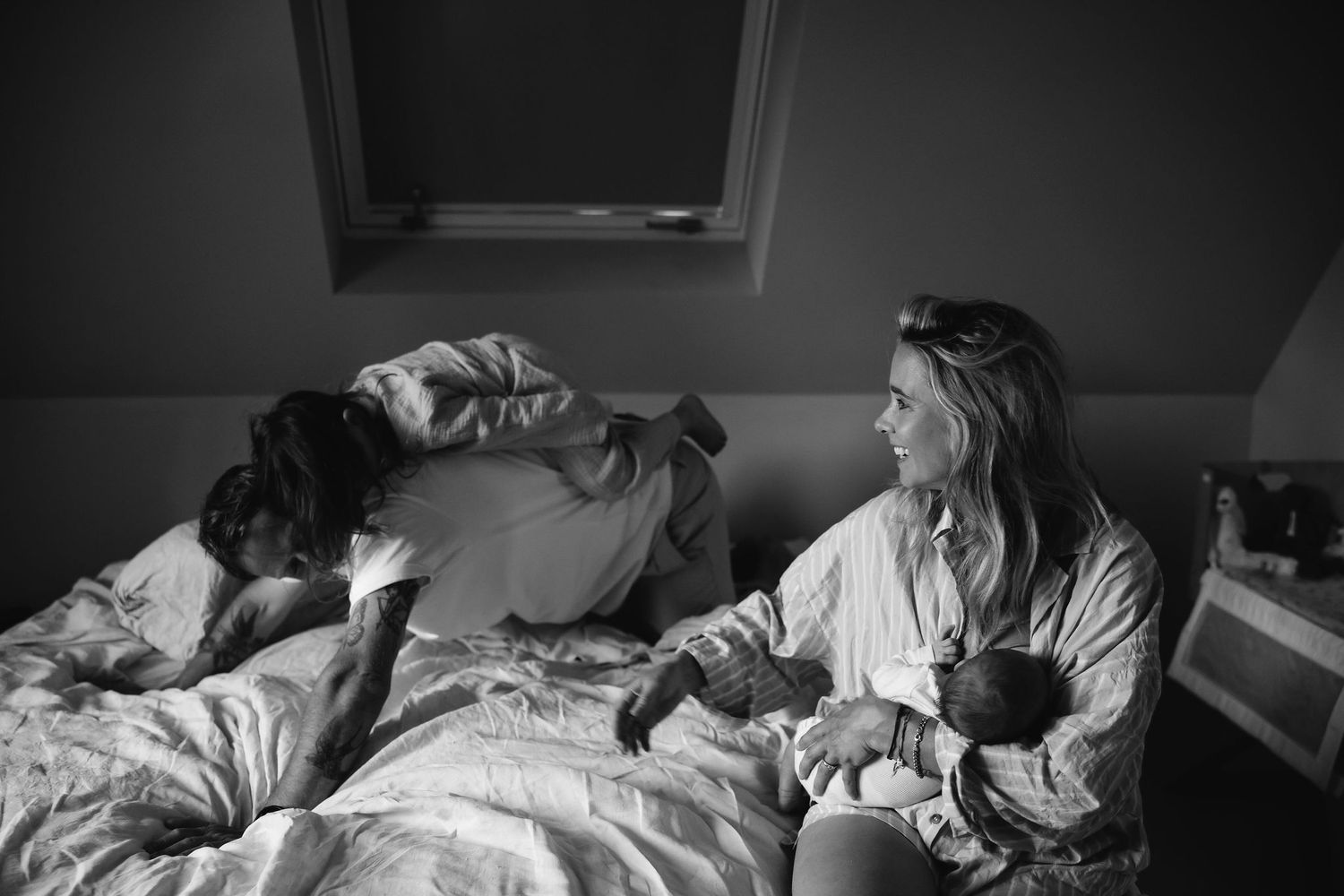 Moody black and white photograph of a bedroom scene with family members on a bed.