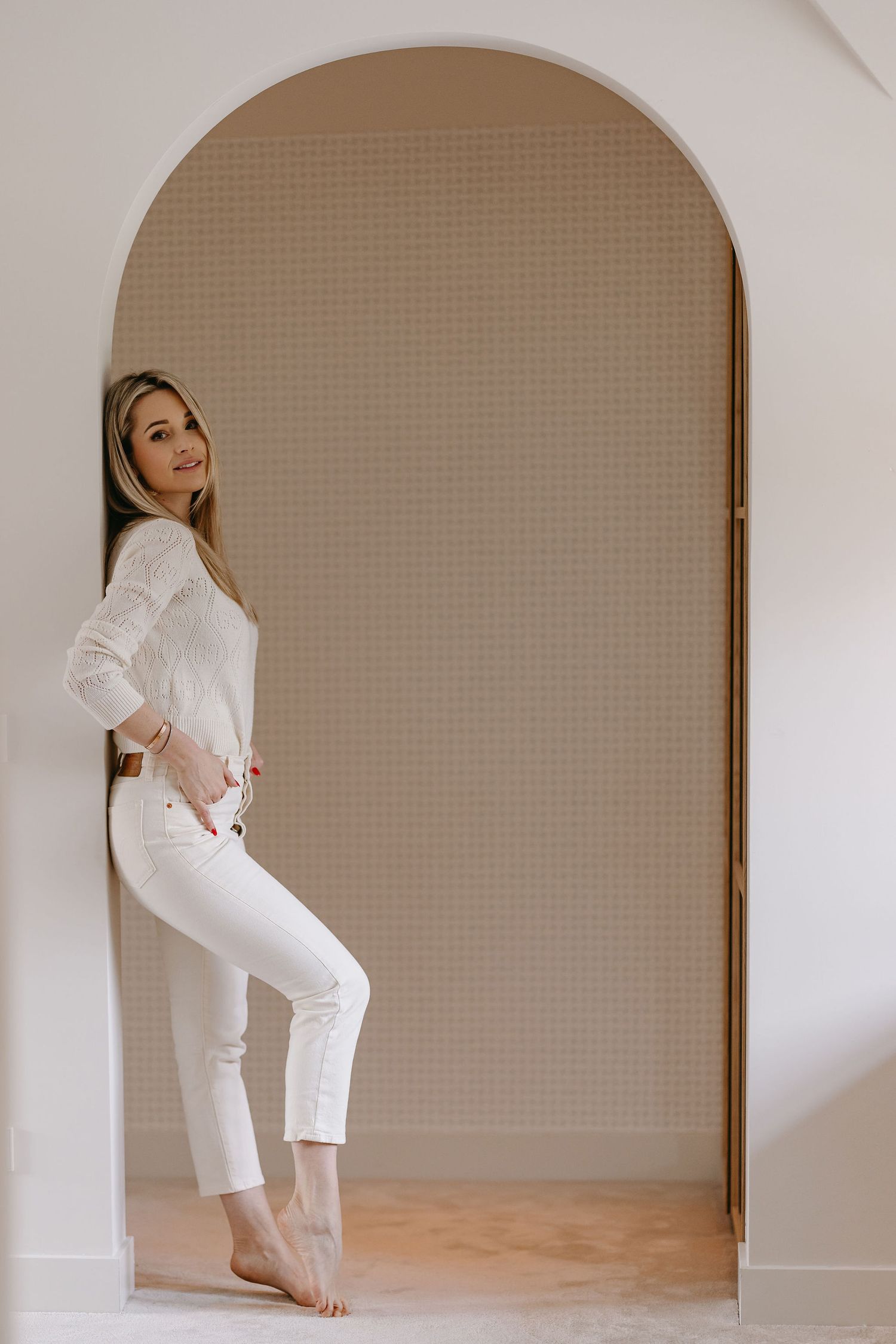 Woman in all-white outfit poses against arched wall during minimalist photo session.