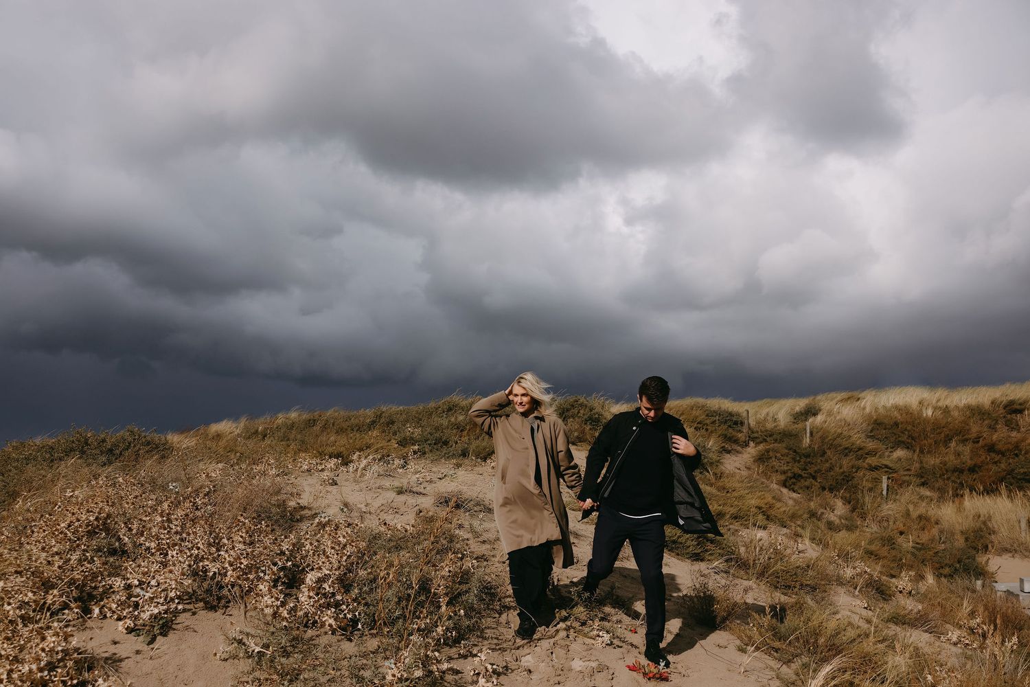A couple walks hand in hand on a hilltop under dramatic storm clouds.