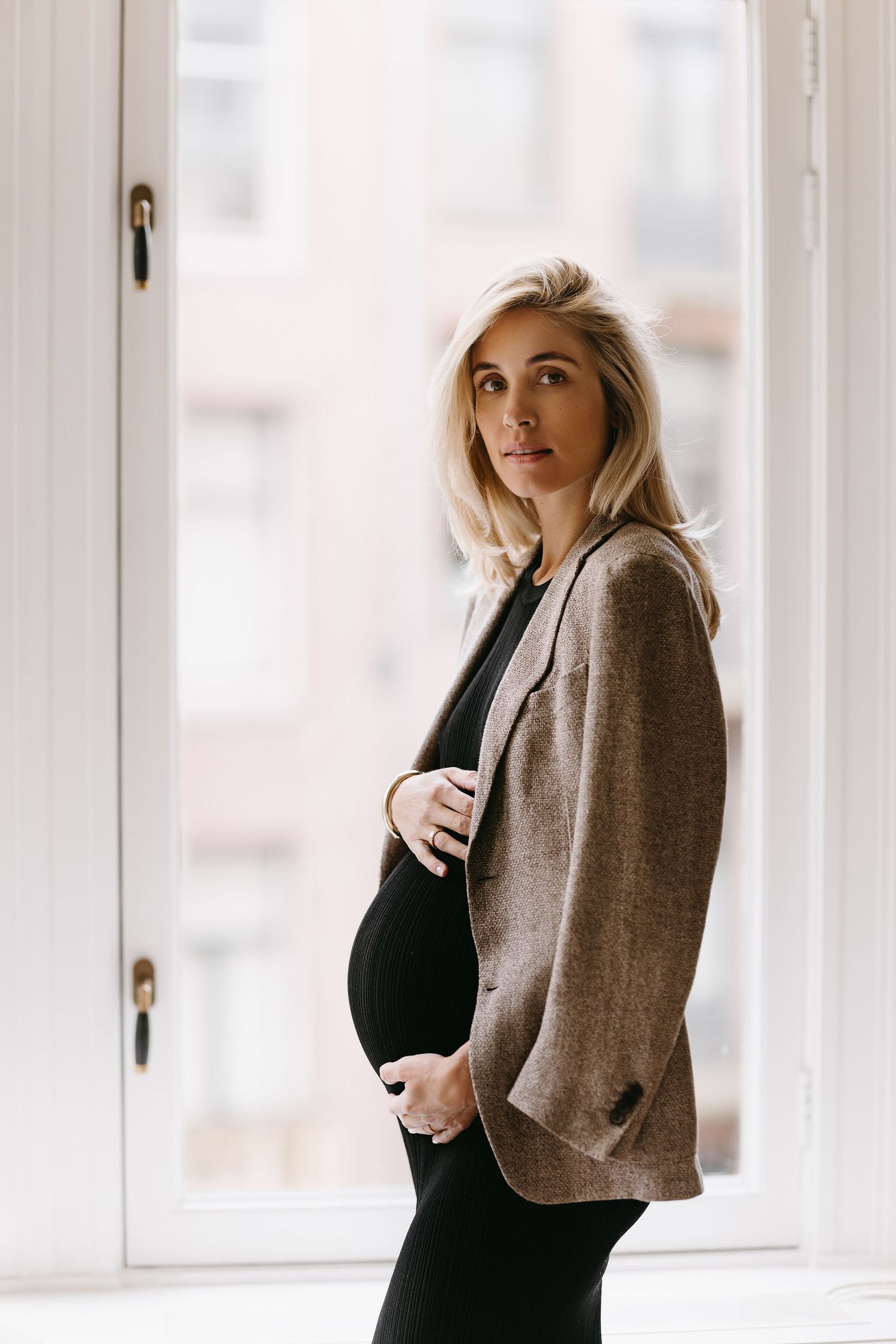 Maternity portrait of person in blazer and black dress by bright window.