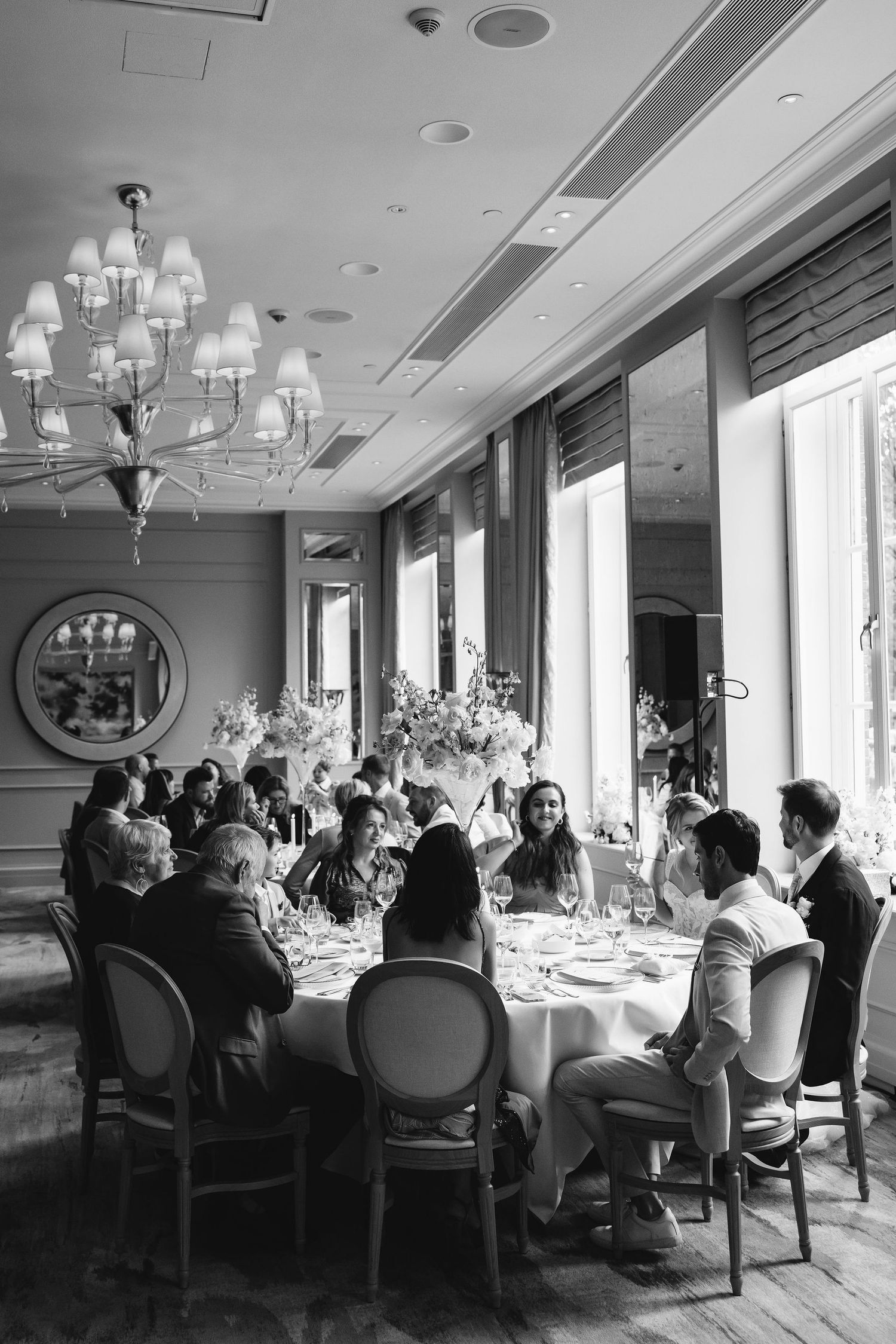 Wedding reception guests dining at elegant round tables under crystal chandeliers.