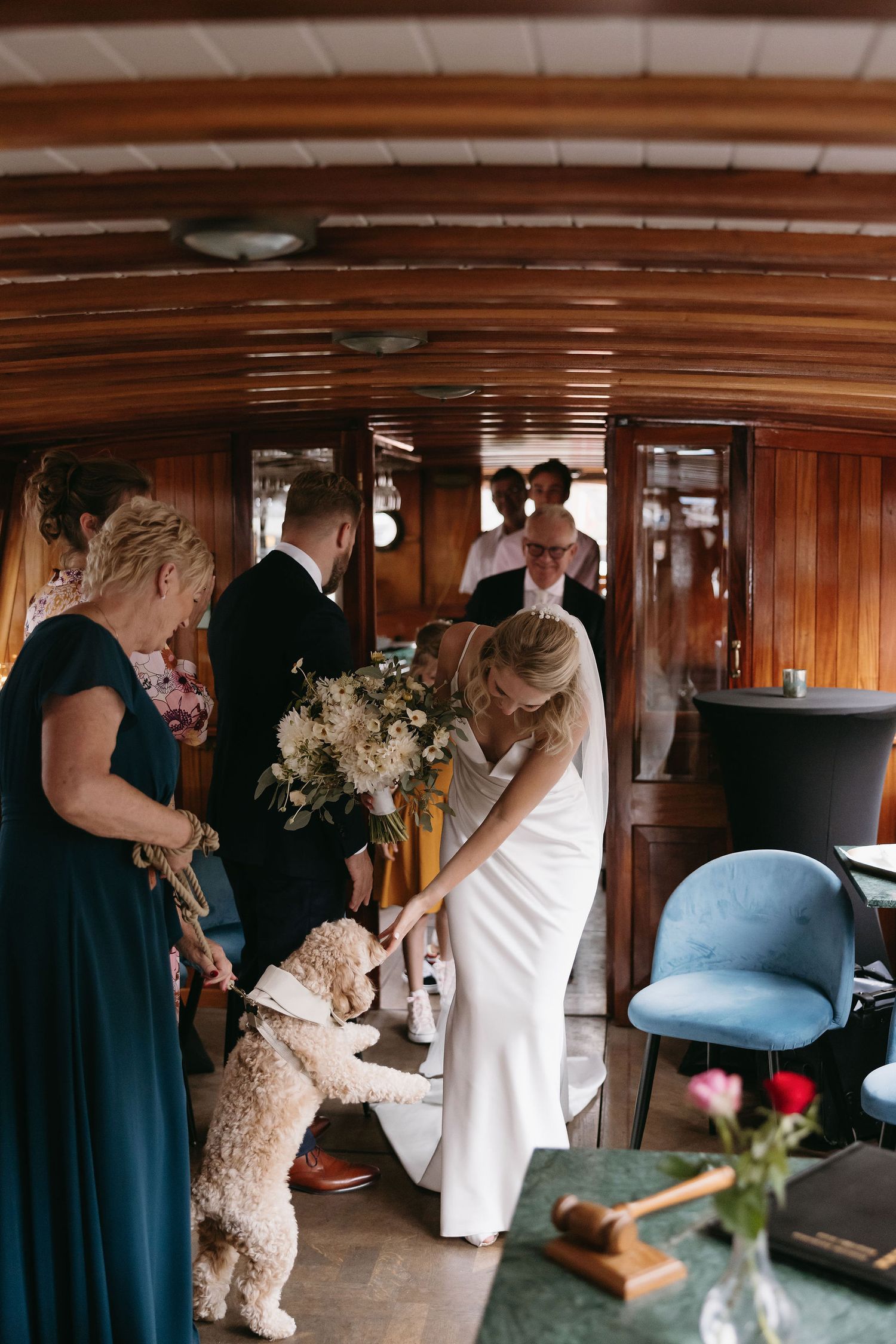 A white fluffy dog greets a bride in a sleek white dress aboard a wooden boat interior with navy-clad guests.