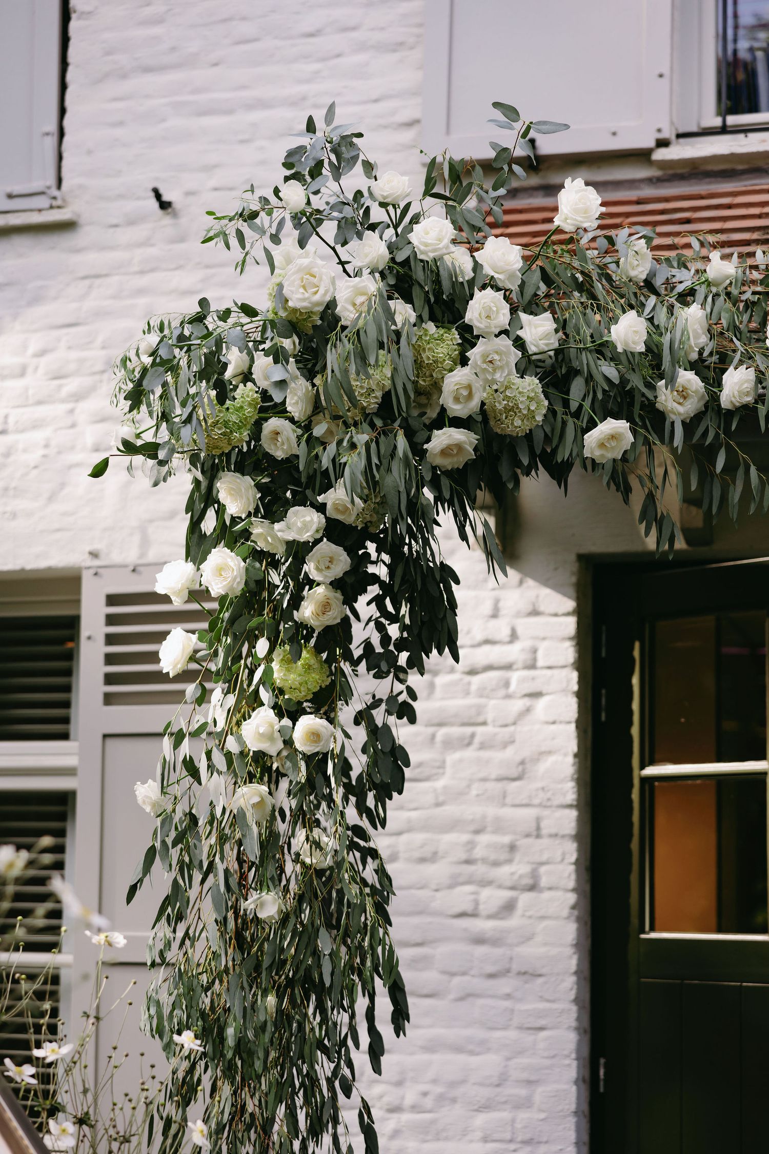 A beautiful floral arch with white roses and eucalyptus decorates a white brick building entrance.