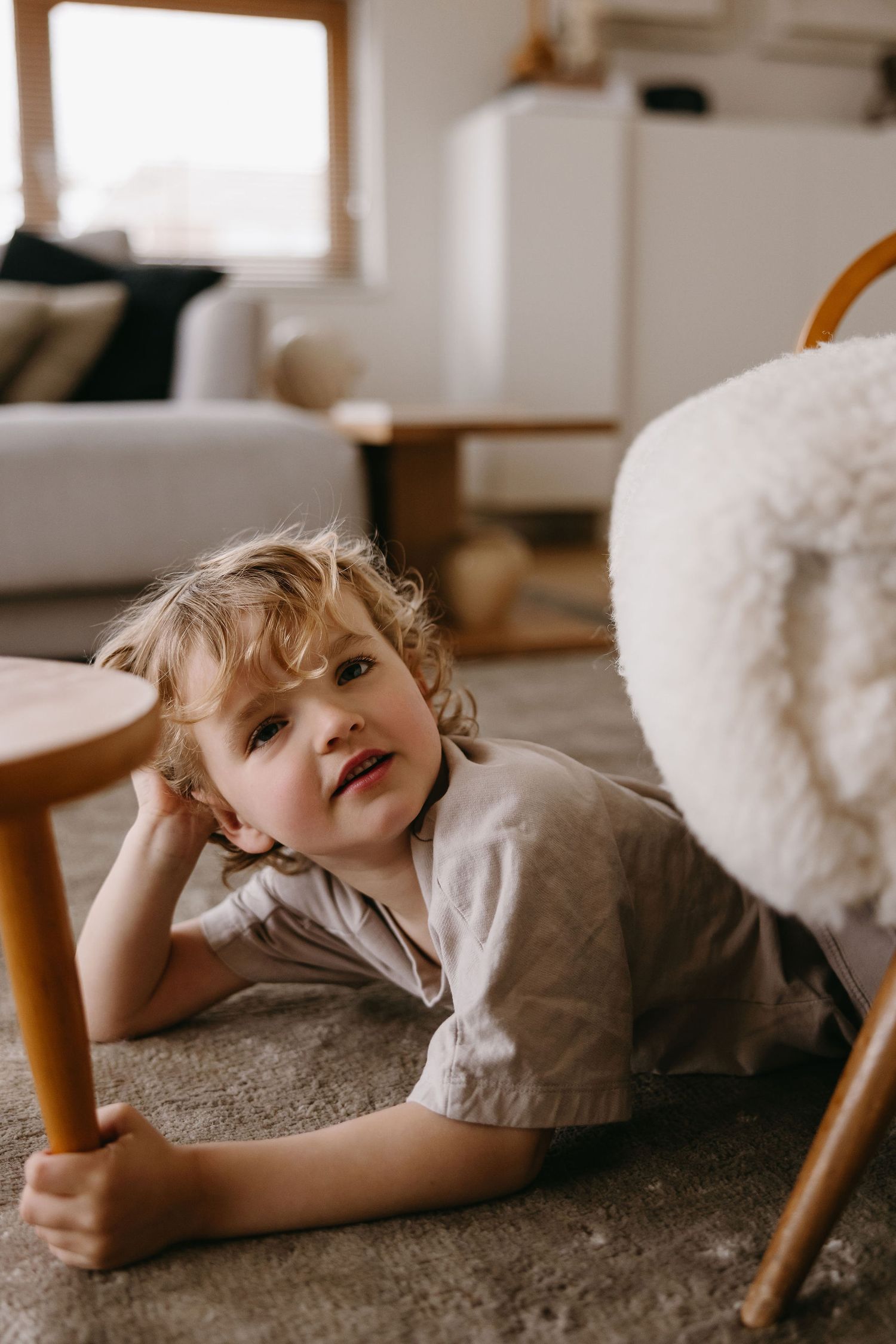 Child relaxing on carpet while holding wooden chair leg in cozy living room.
