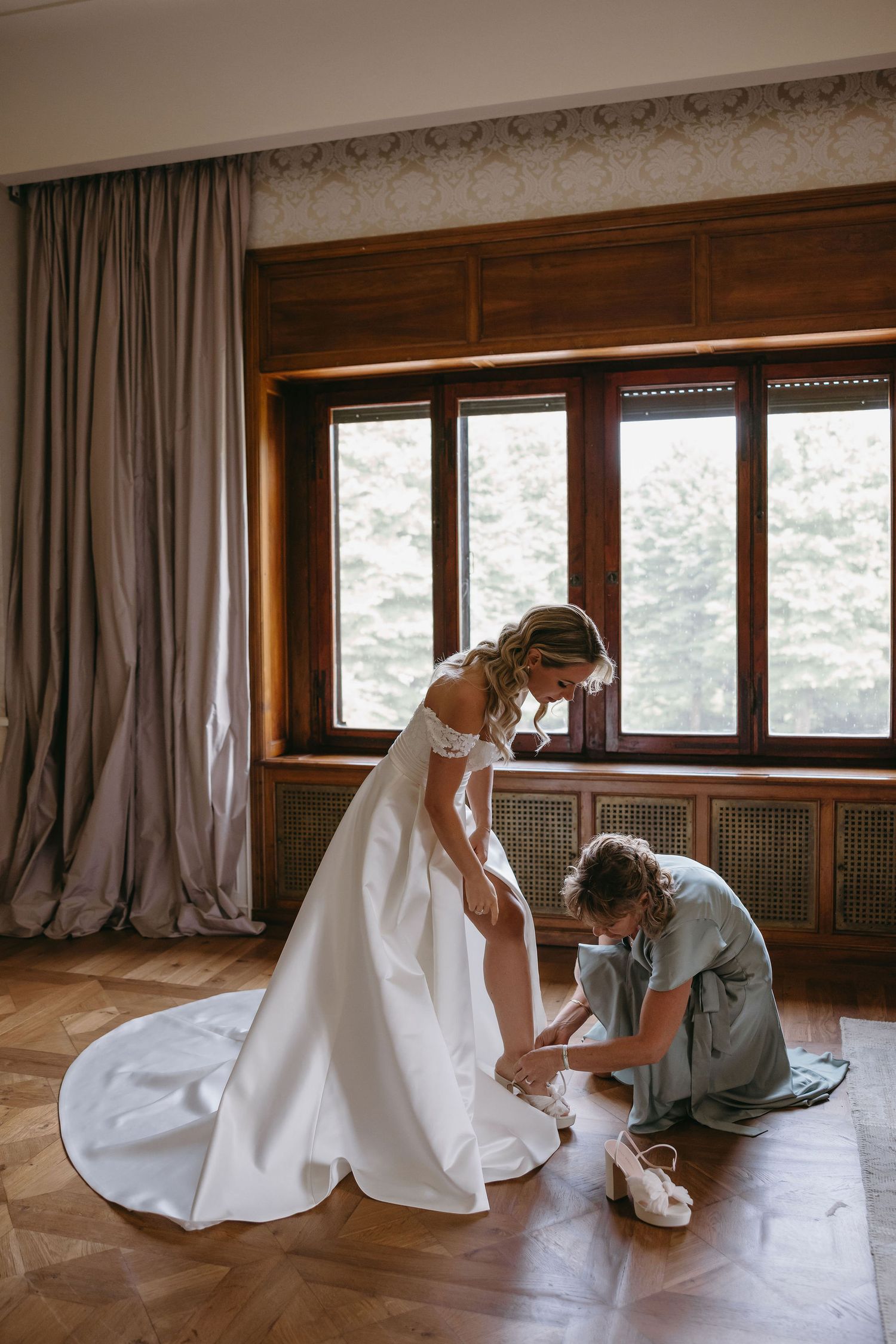 Getting ready moment as someone helps adjust a wedding shoe in a sunlit room.