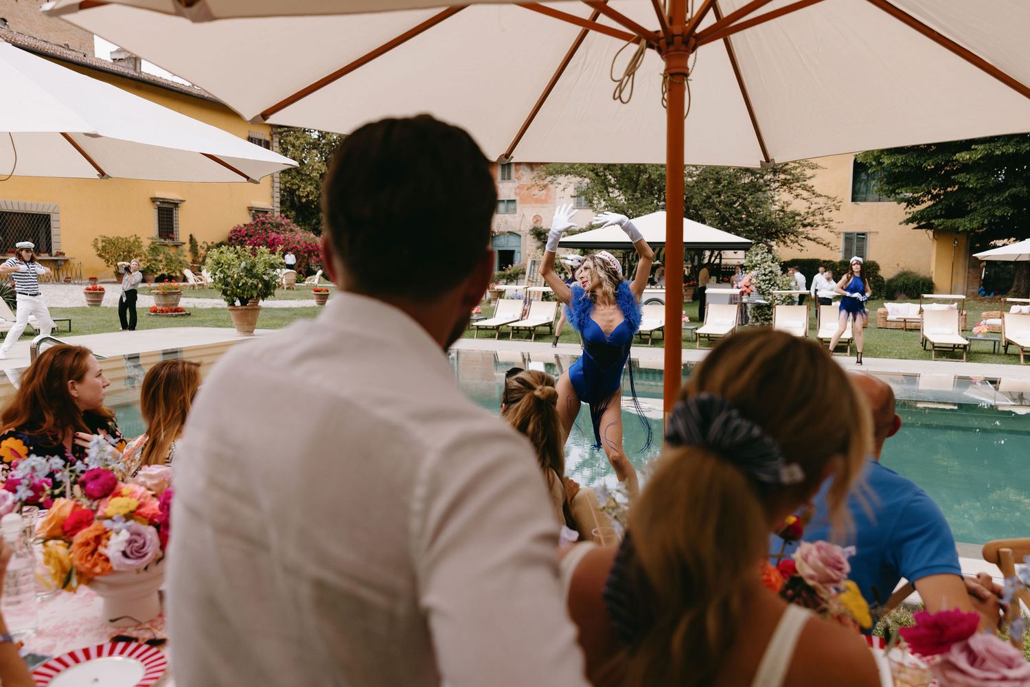 Outdoor wedding reception by the pool with guests gathered under white umbrellas.
