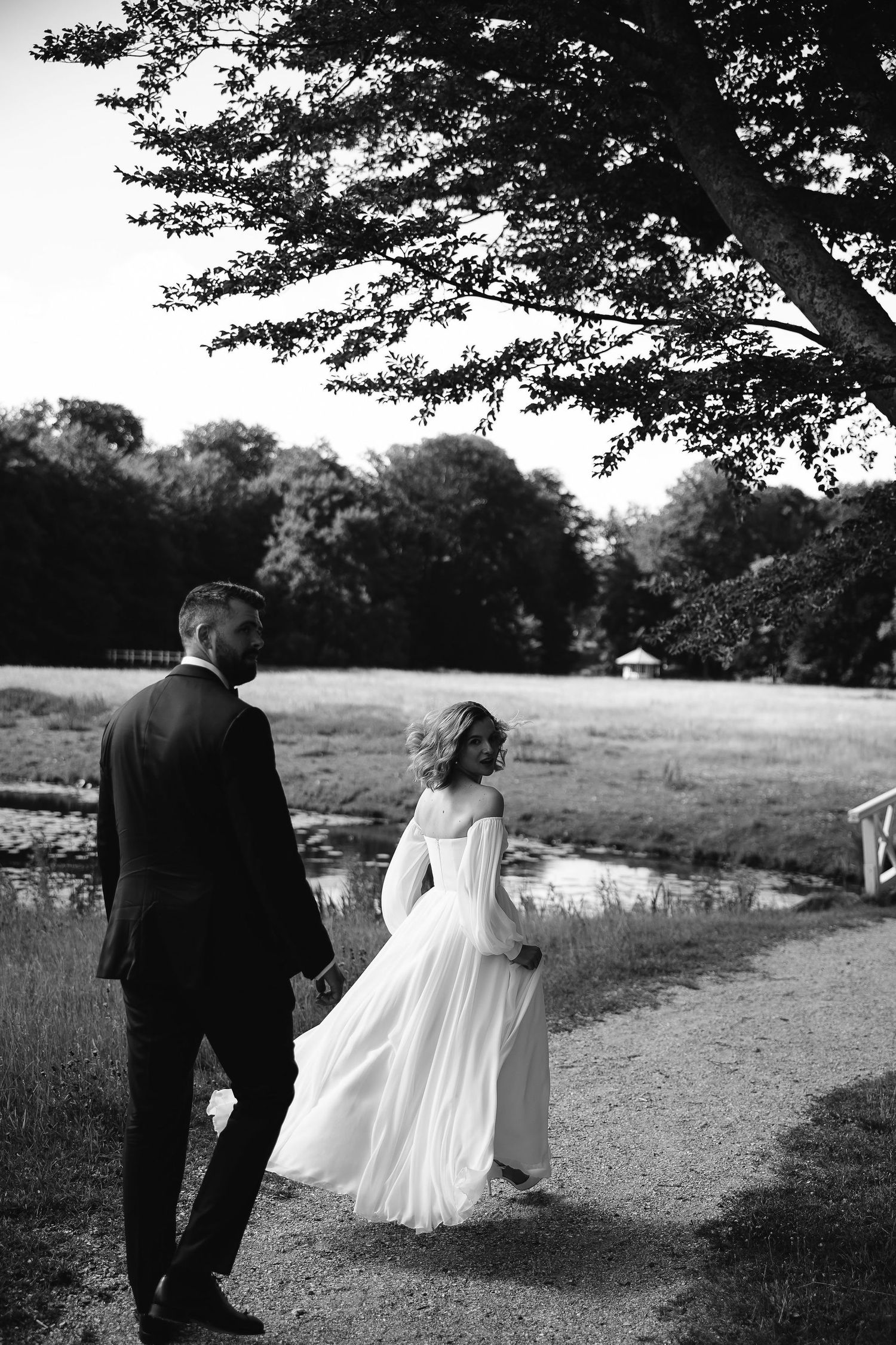 A wedding couple walks along a tree-lined path in black and white photography.
