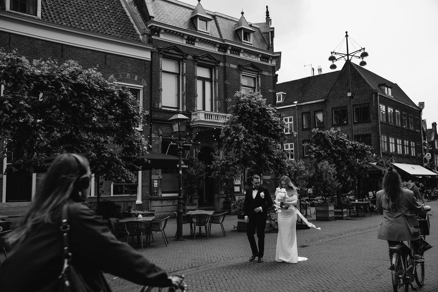 Wedding couple walking down a historic street while cyclists pass by in black and white.