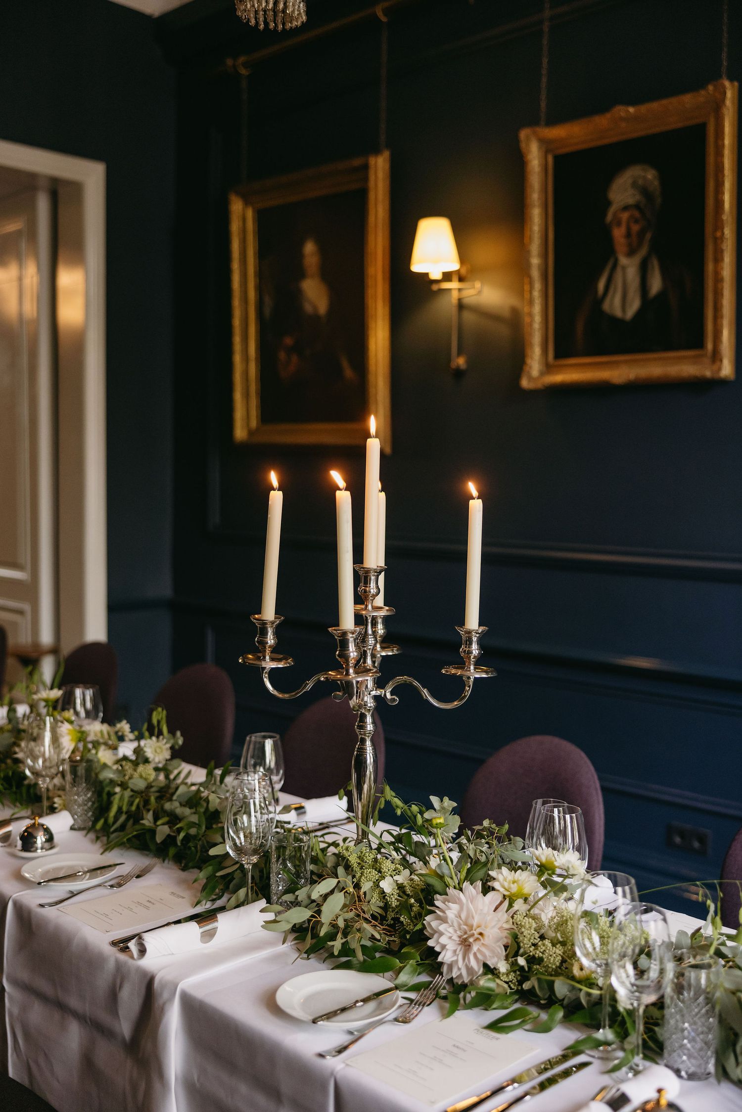 Intimate wedding reception table with silver candelabras and greenery runner against navy blue walls.