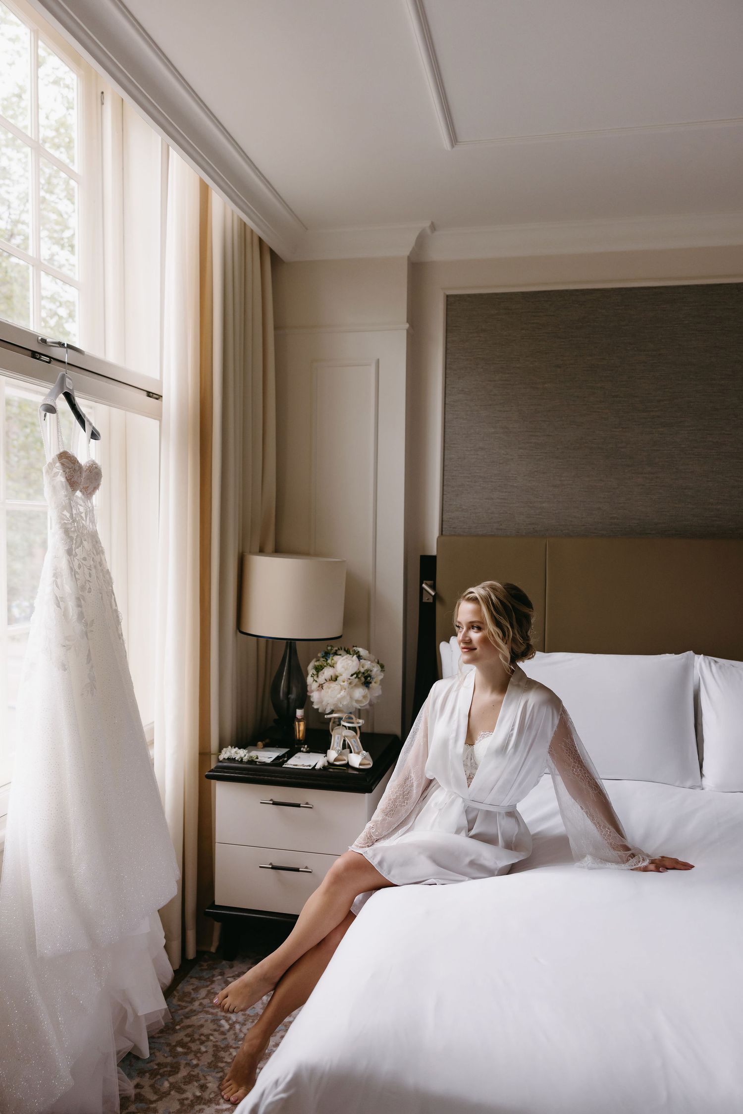 Wedding dress hanging by window in elegant hotel room with bride on bed.
