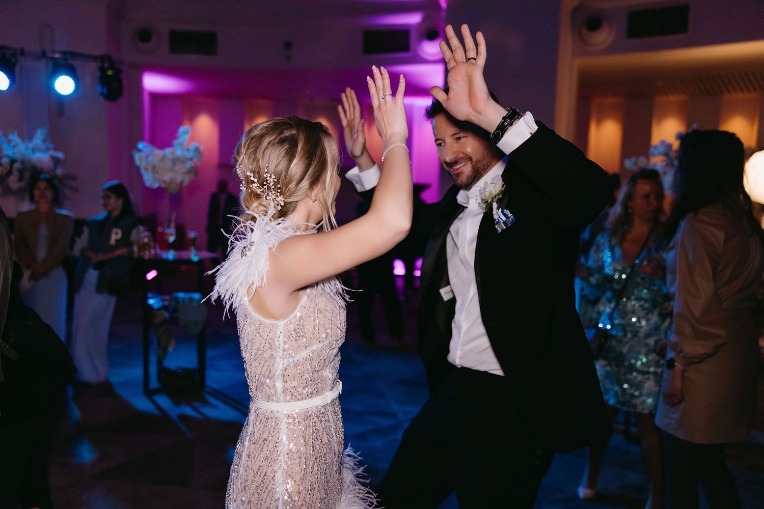 Couple dancing energetically at wedding reception with colorful lighting.