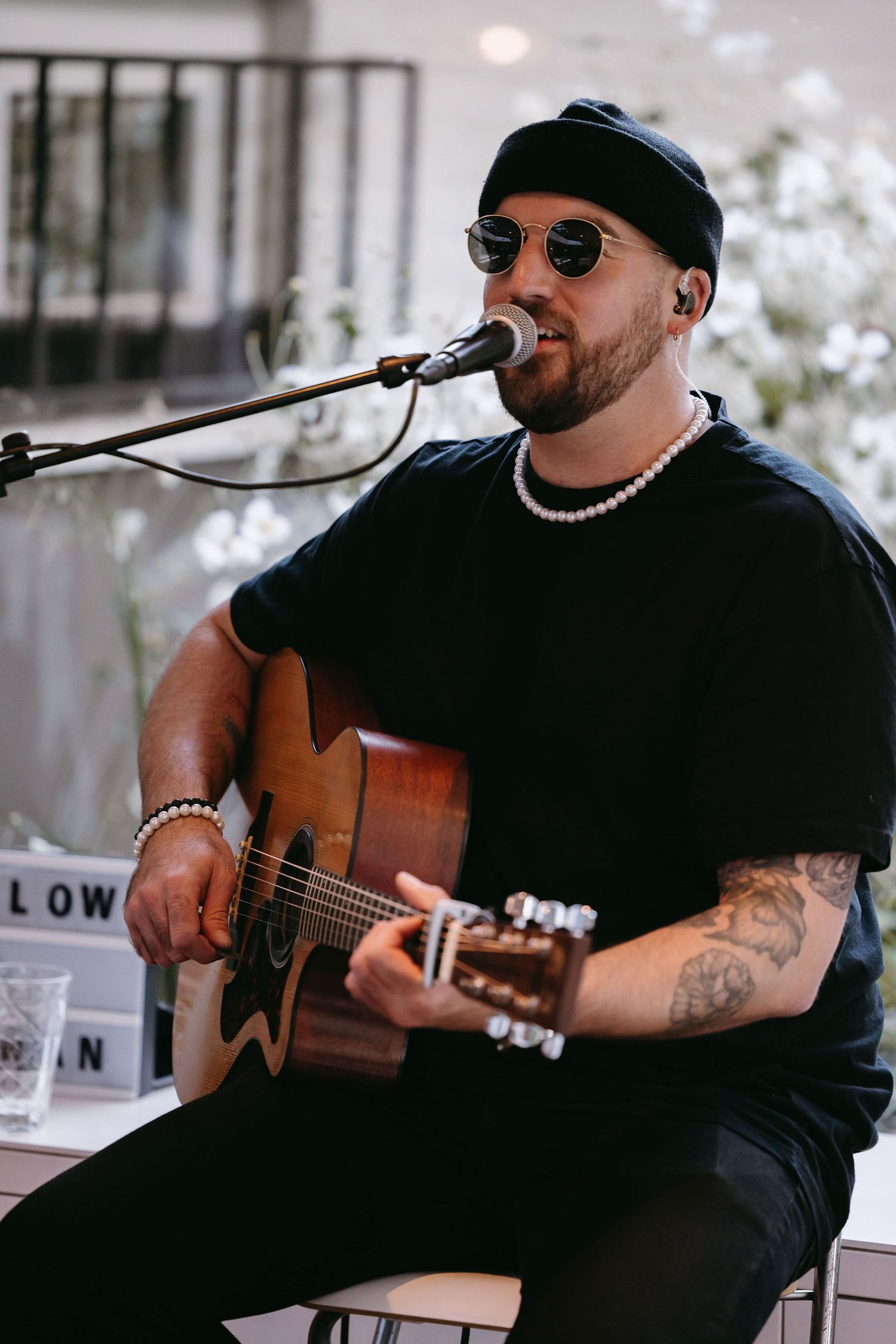 Musician in sunglasses and black attire performs with acoustic guitar at outdoor venue.