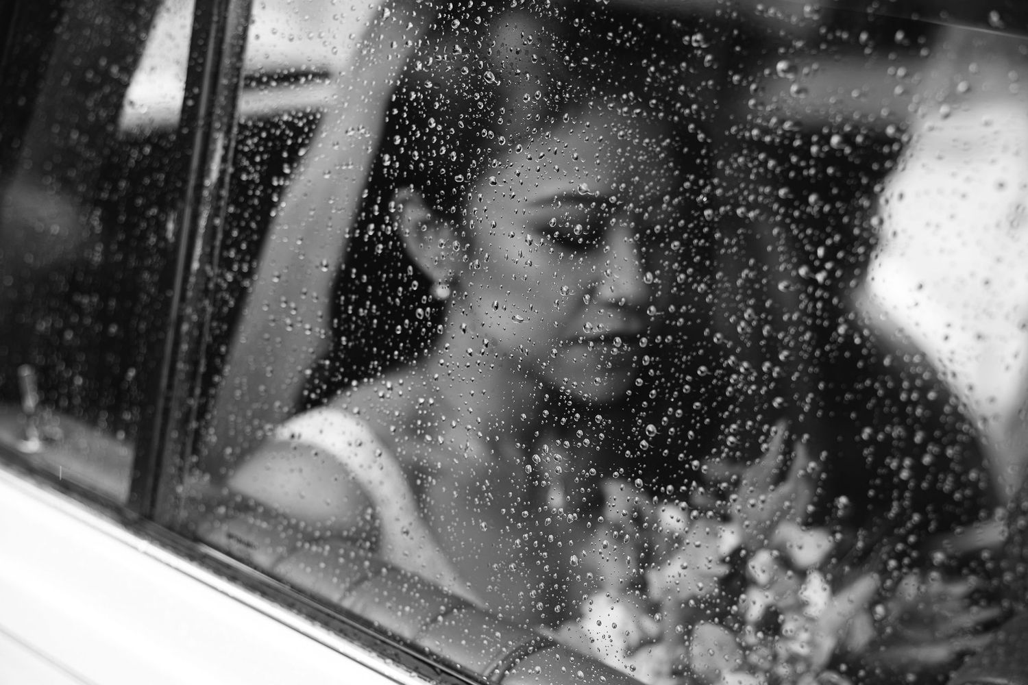 A black and white photograph through a rainy window shows a person gazing out while holding flowers.