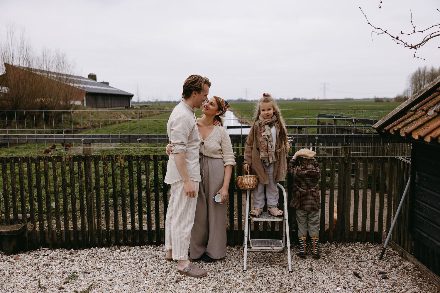 Three people stand together in casual attire on a rustic deck overlooking pastoral farmland on an overcast day.