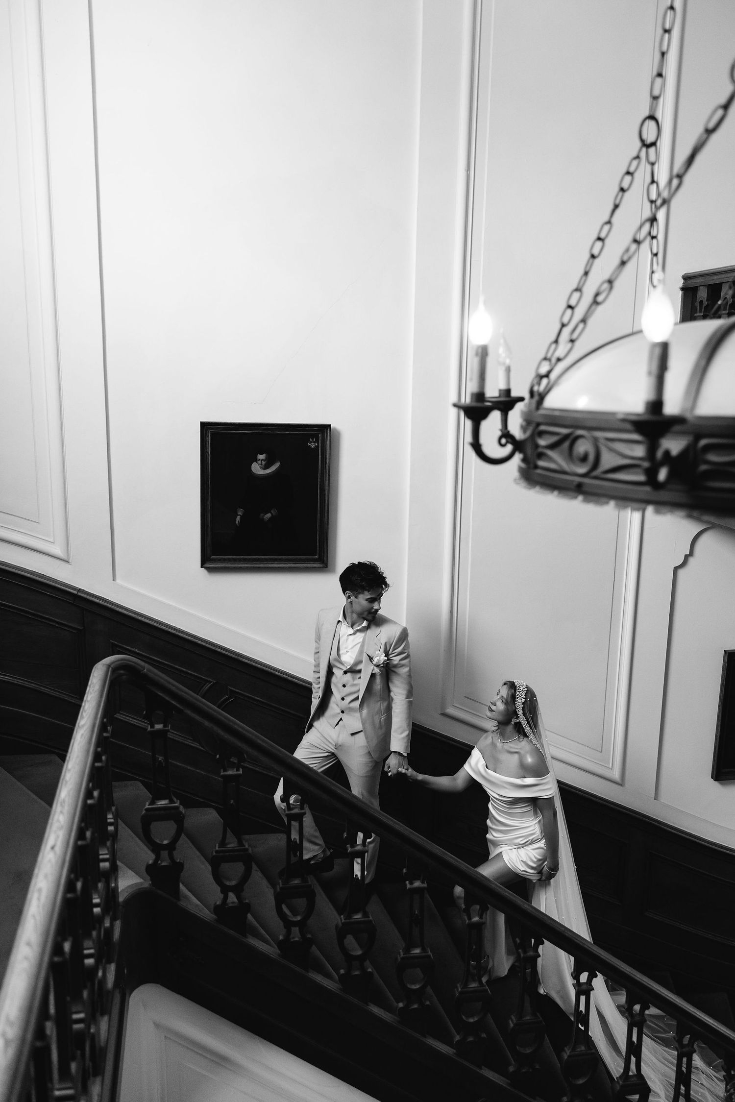 Black and white photo of a winding staircase in an elegant vintage interior with a chandelier and wall panels.