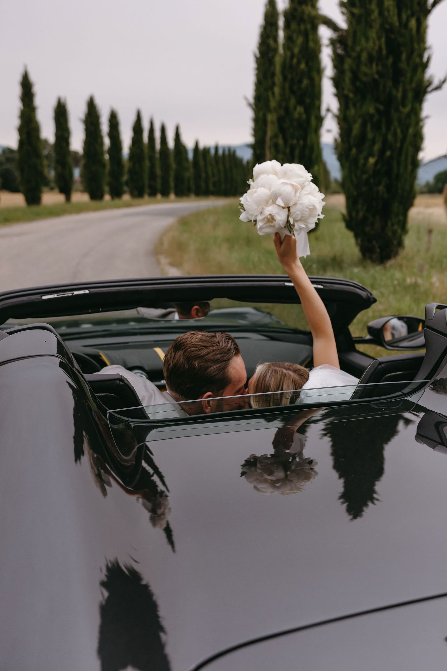 White orchid bouquet held over a classic dark convertible car with cypress trees in the background.